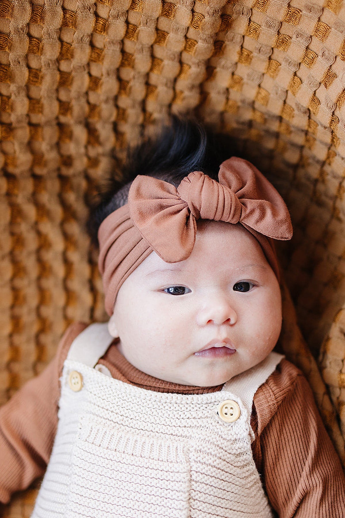 A baby wearing a rust-colored head wrap and cream knit overalls on a textured brown blanket.