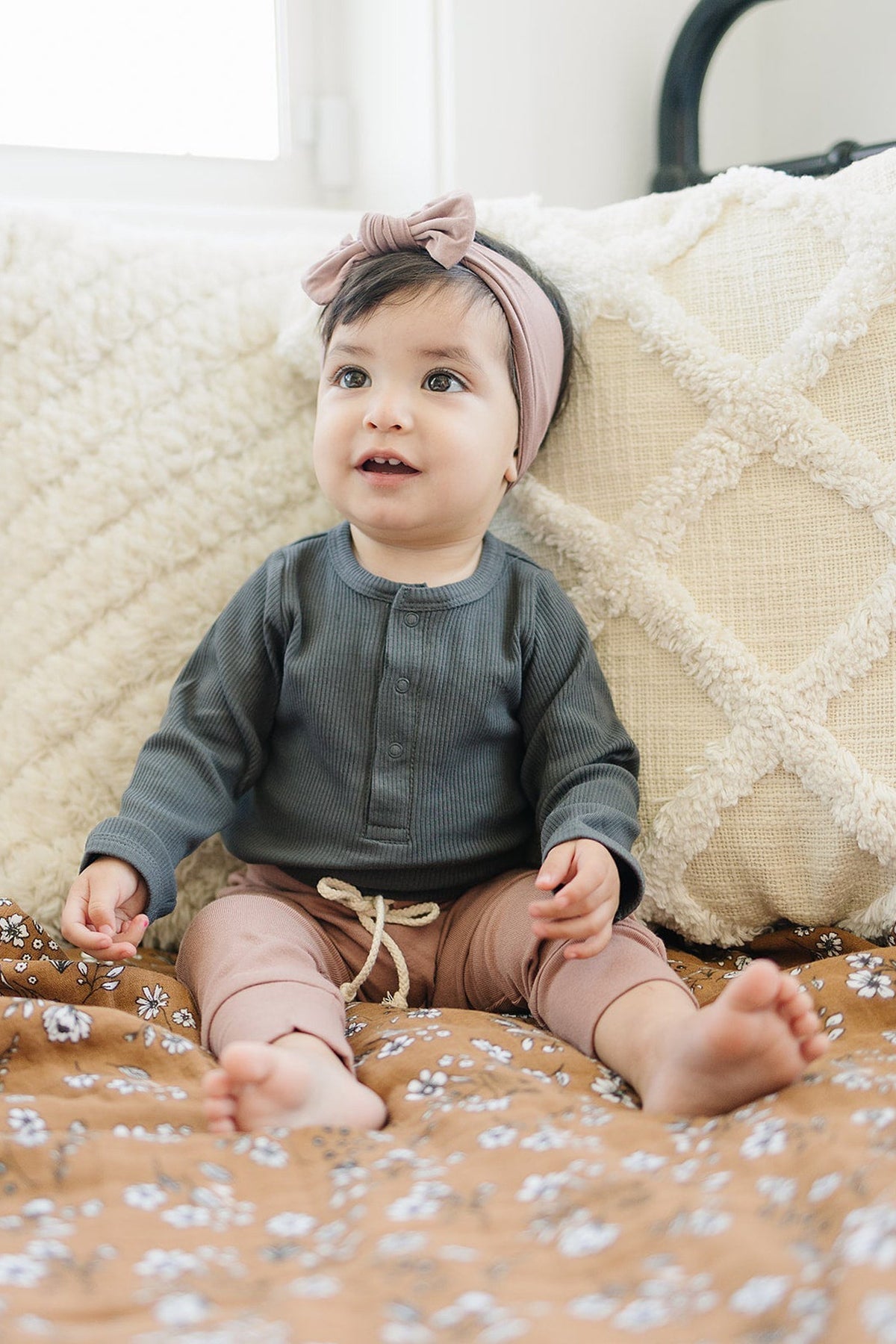 A baby wearing a charcoal long-sleeve top and pink headband sits on a floral blanket with a textured pillow.