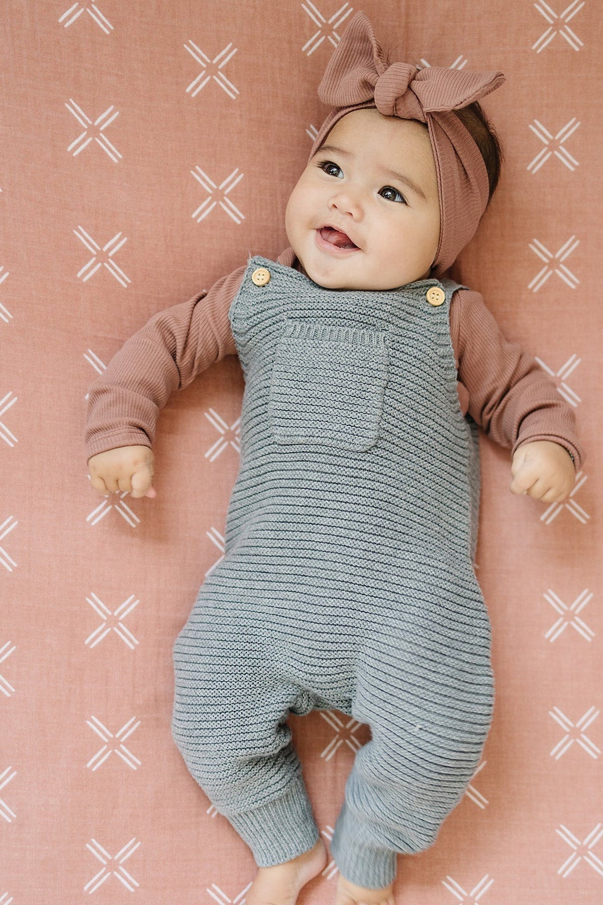 A baby in a knitted blue romper and headband lies on a pink patterned muslin crib sheet.