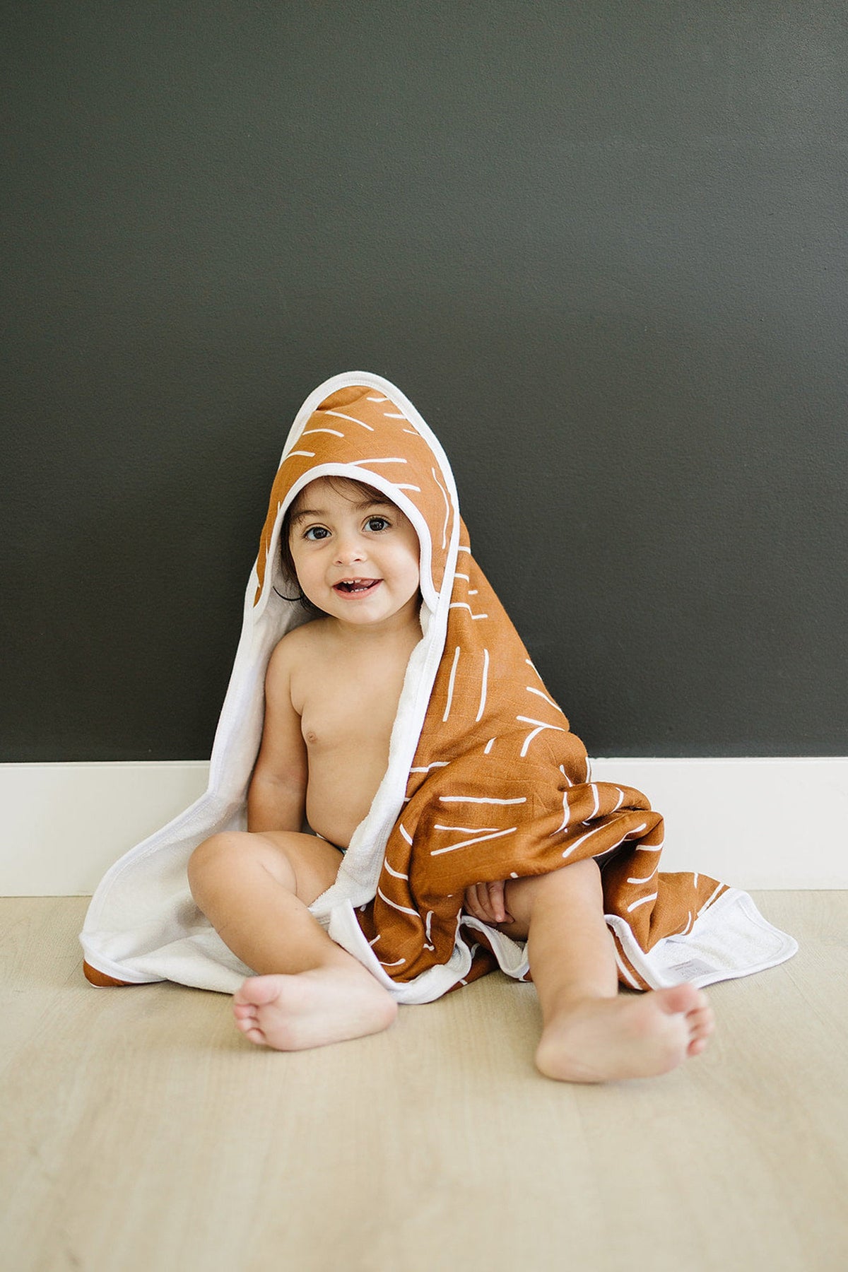 A baby sits wrapped in a mustard-colored hooded towel with white patterns against a dark wall.