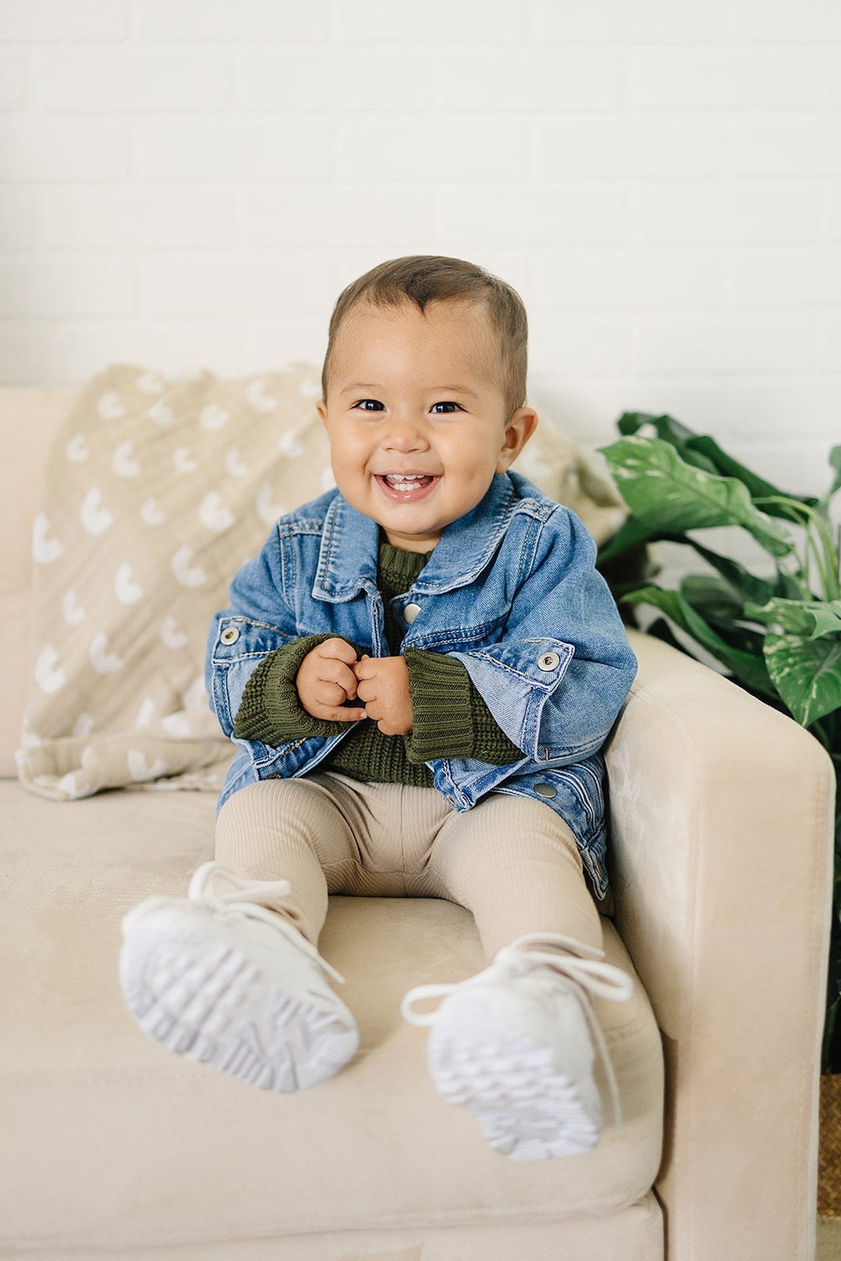 A smiling baby wearing a blue denim jacket and beige leggings sits on a cream sofa with a plant nearby.