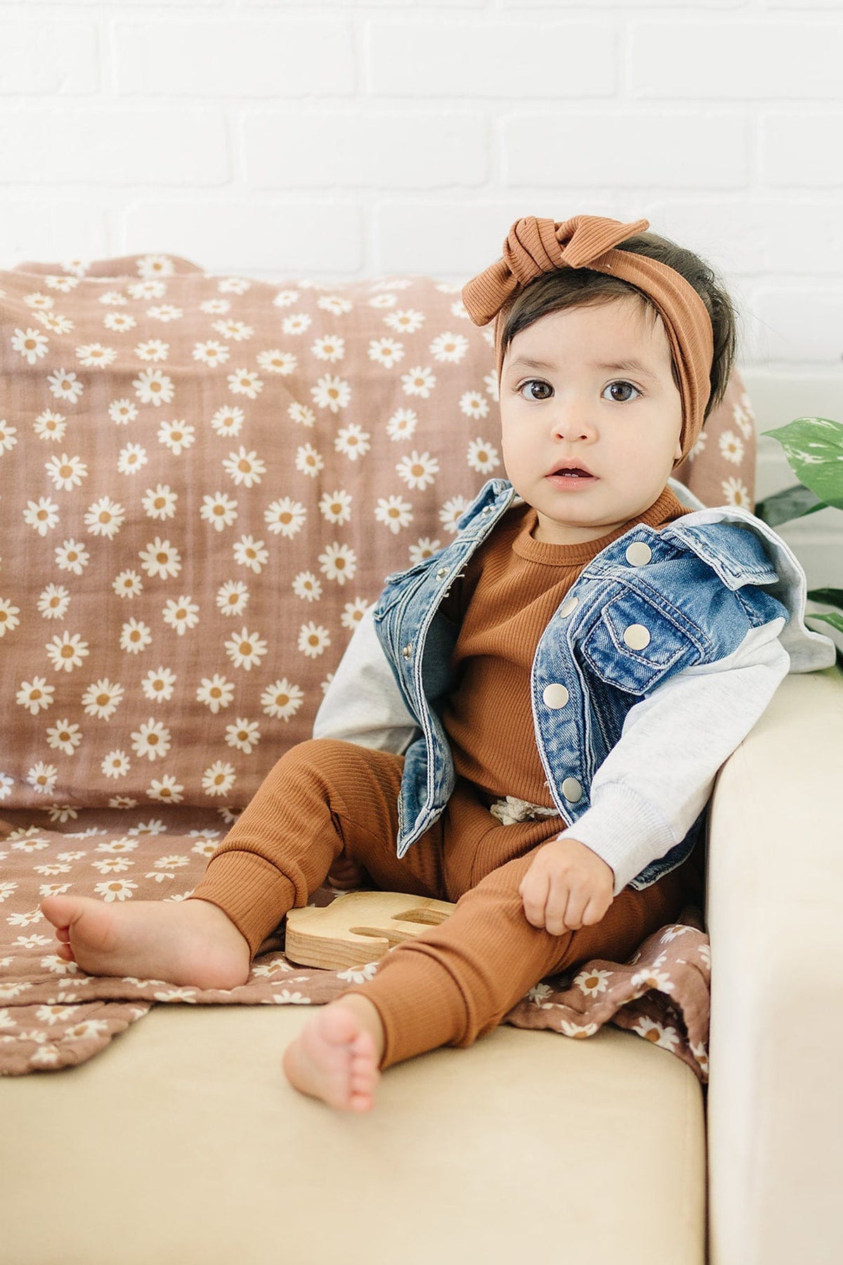 Baby in a denim and grey hooded jacket sits on a floral-patterned blanket, wearing a brown outfit and headband.
