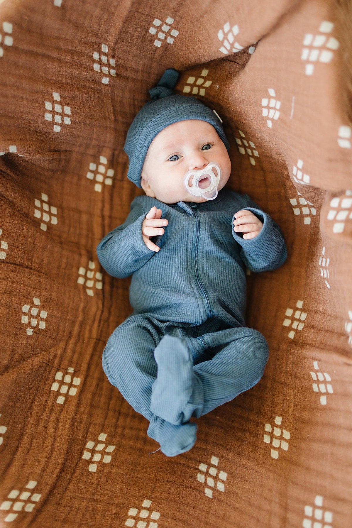 Baby wearing a navy ribbed outfit and knot hat, lying on a patterned brown blanket, holding a pacifier.