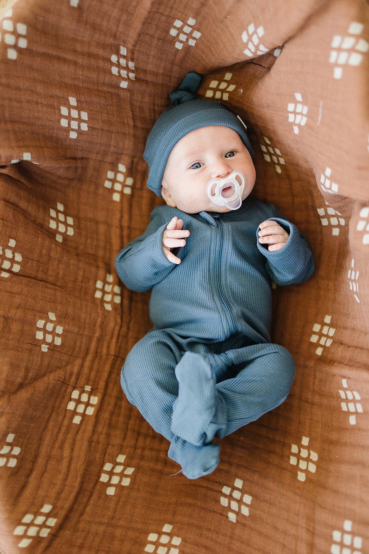 Baby in navy ribbed footed pajama with a pacifier, lying on a patterned brown blanket.
