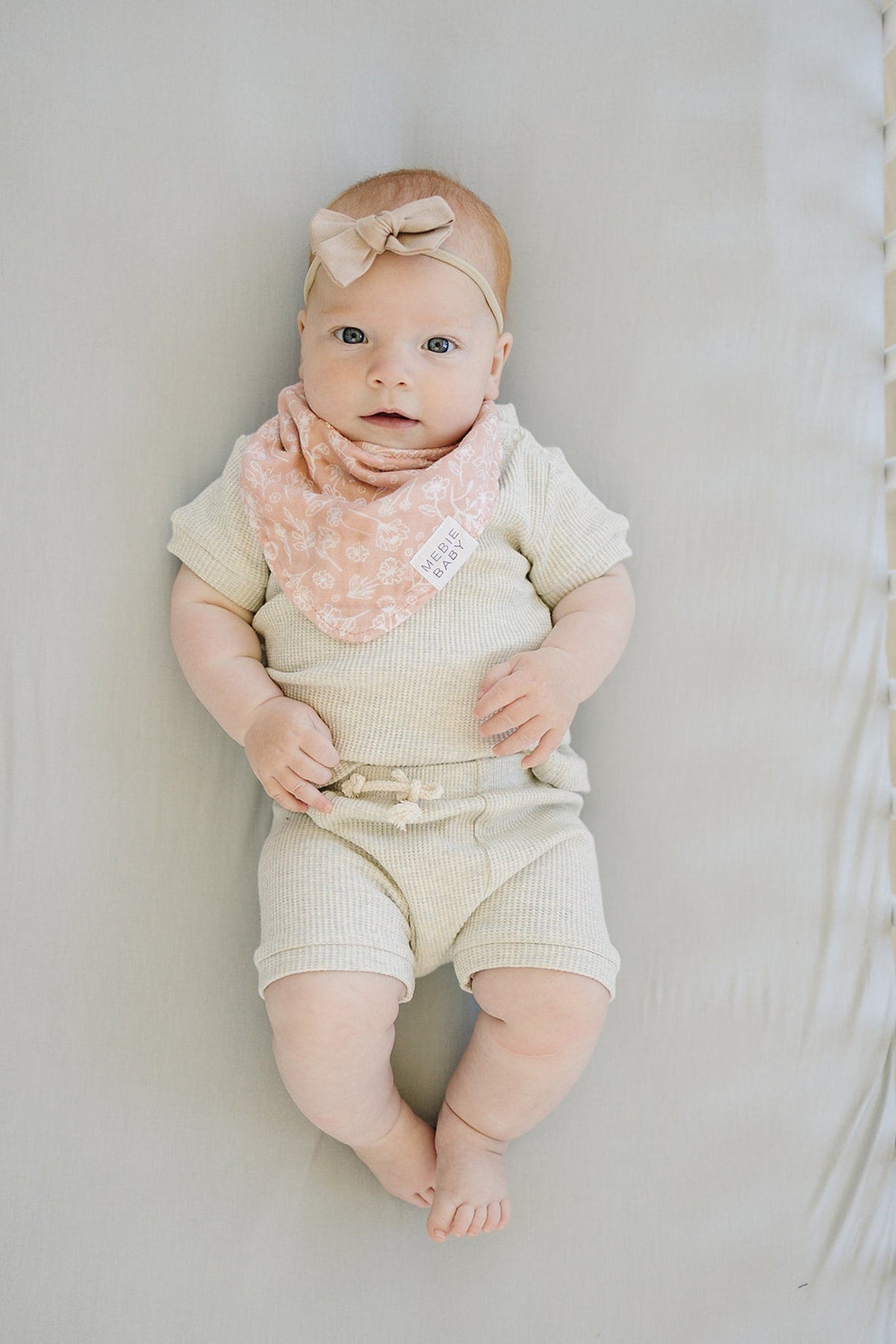 A baby wearing a beige outfit and pink bib with a bow headband lies on a light gray sheet.