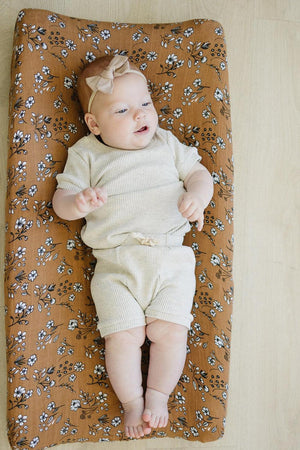 A baby lies on a brown floral muslin changing pad cover, wearing a cream outfit and headband.