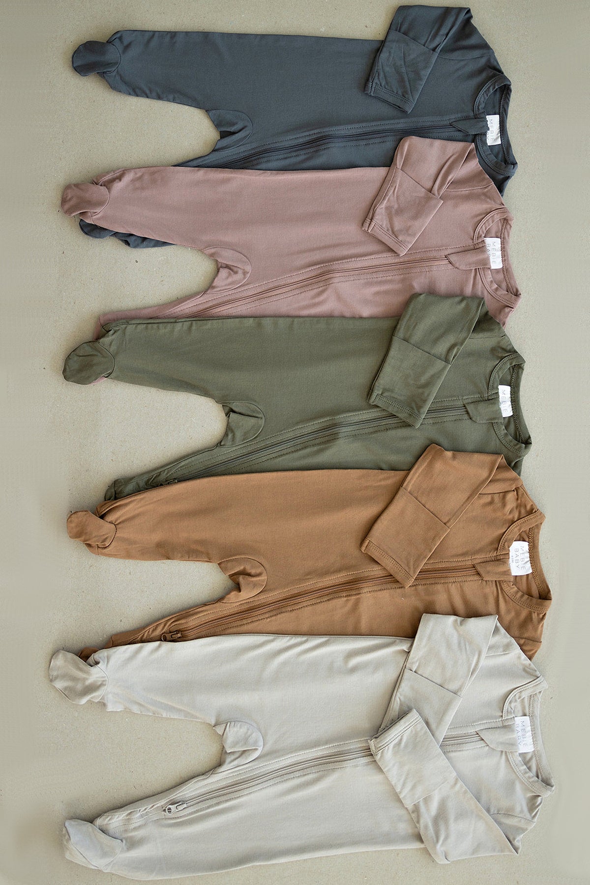 Five footed baby pajamas in various colors, including gray, mauve, olive, brown, and cream, displayed on a beige background.
