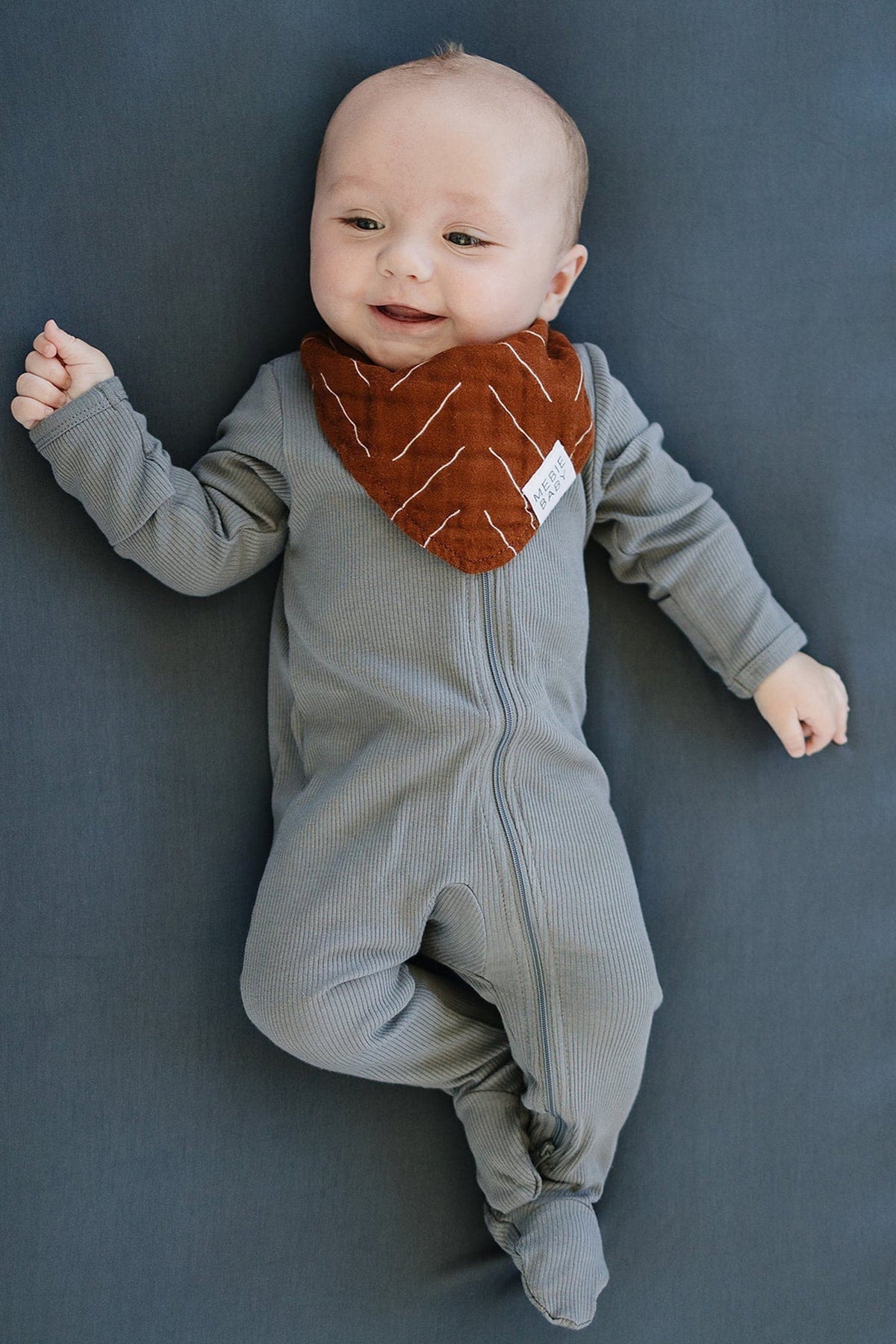 Smiling baby wearing a gray onesie and brown bib lies on a charcoal crib sheet.