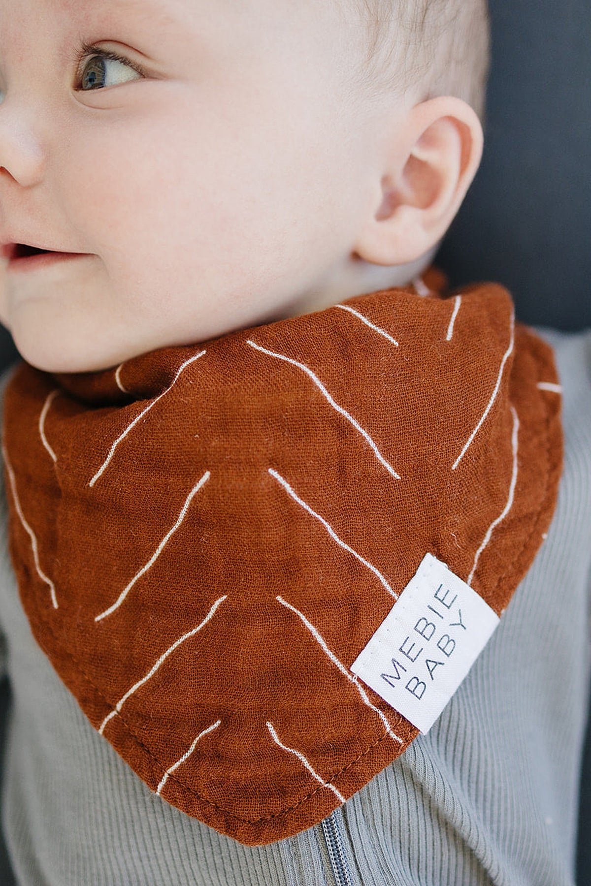 A baby wearing a rust-colored Mebie Baby muslin bib with white geometric lines, showcasing soft fabric texture.