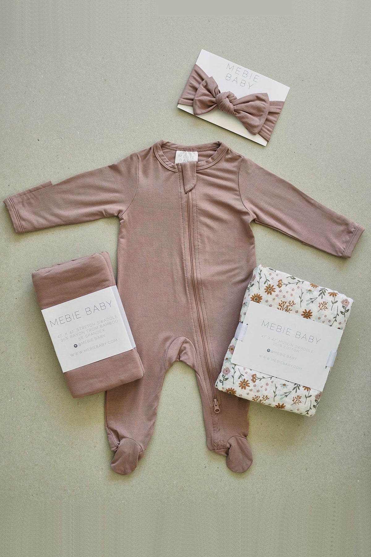 Dusty rose footed pajama with a zipper, matching headband, and two swaddles from Mebie Baby displayed on a surface.