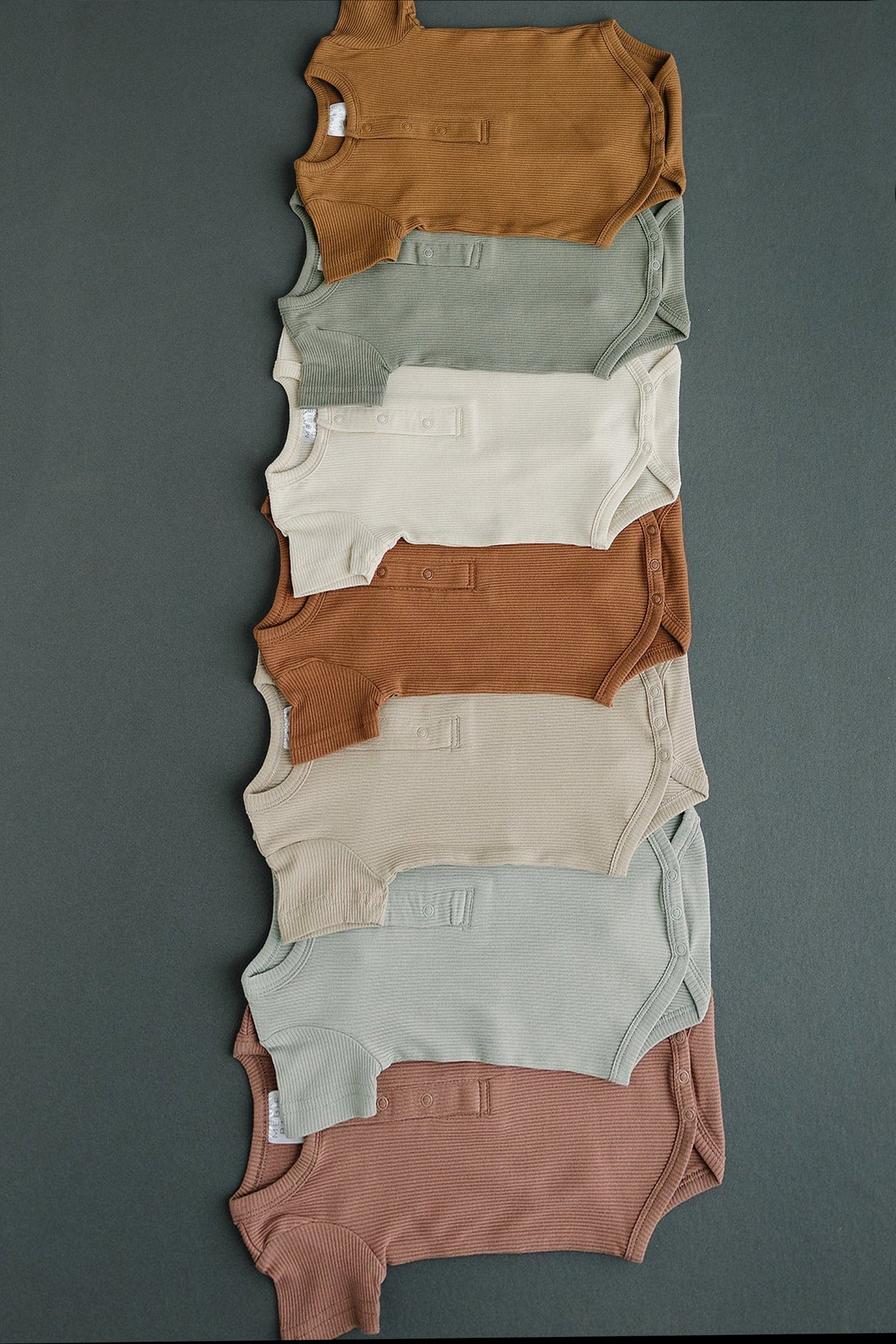 Seven ribbed baby bodysuits in earthy tones are neatly arranged on a dark surface, showcasing their organic cotton fabric.