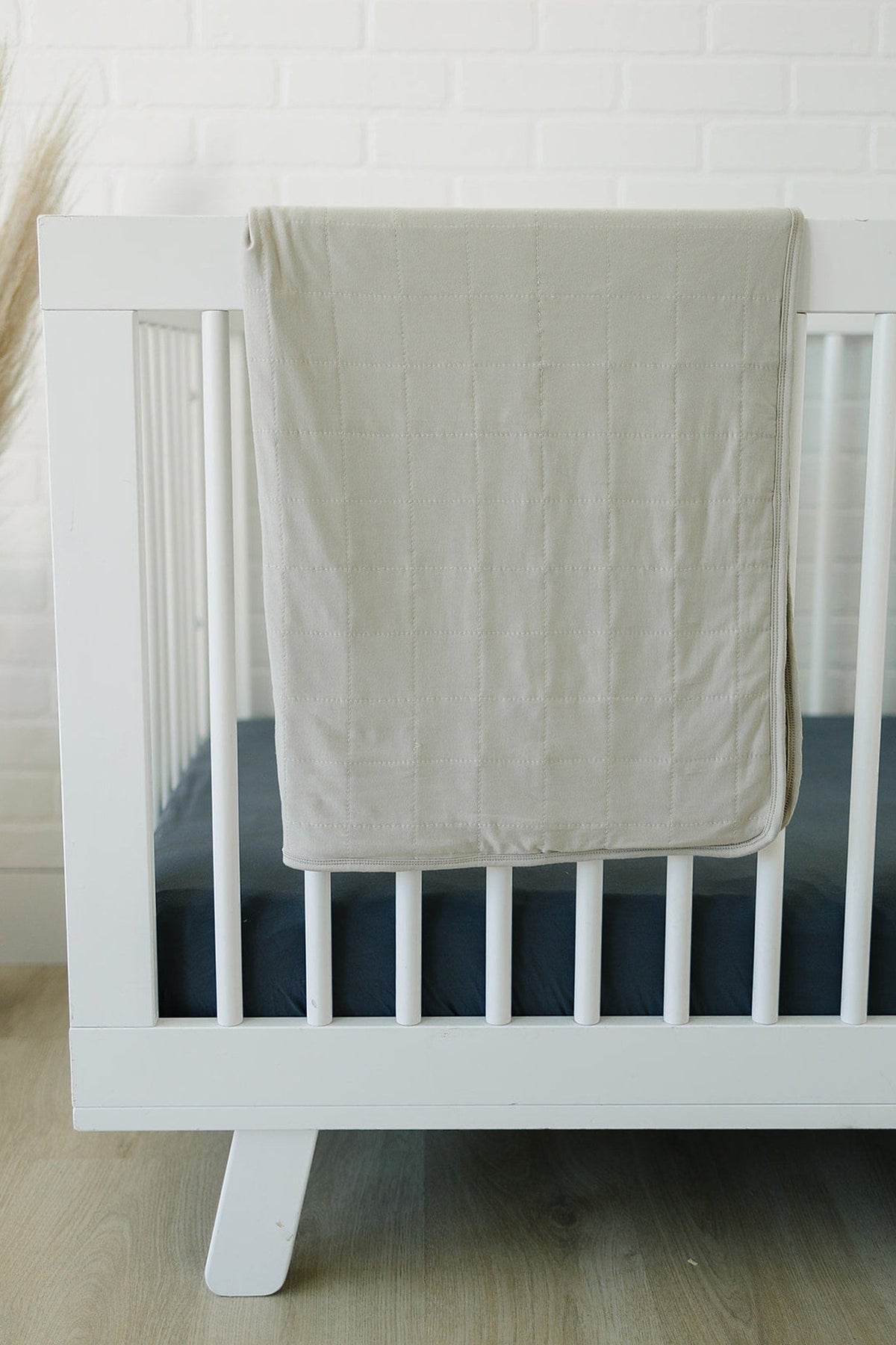 A white wooden crib with a gray quilted blanket draped over the side, on a wooden floor.