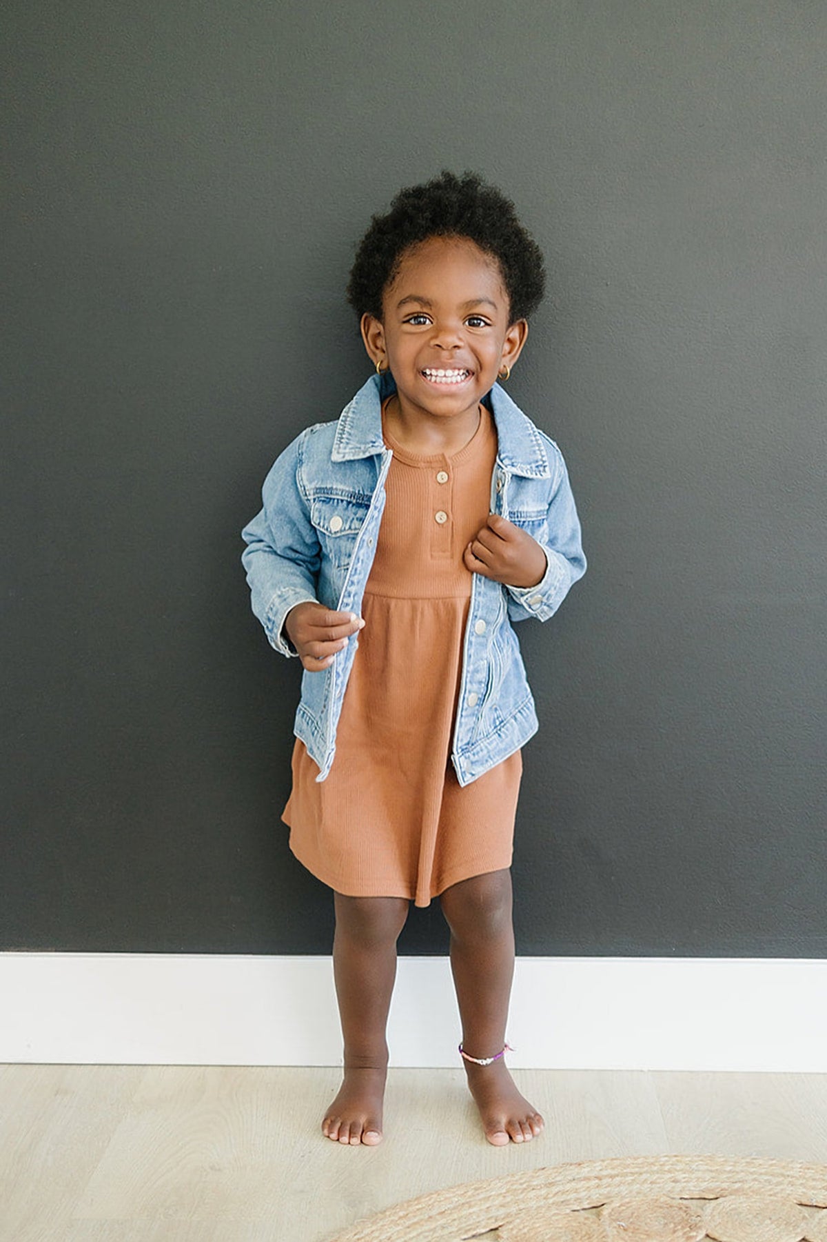 A child smiling, wearing a light blue denim jacket over a brown dress, stands against a dark background.