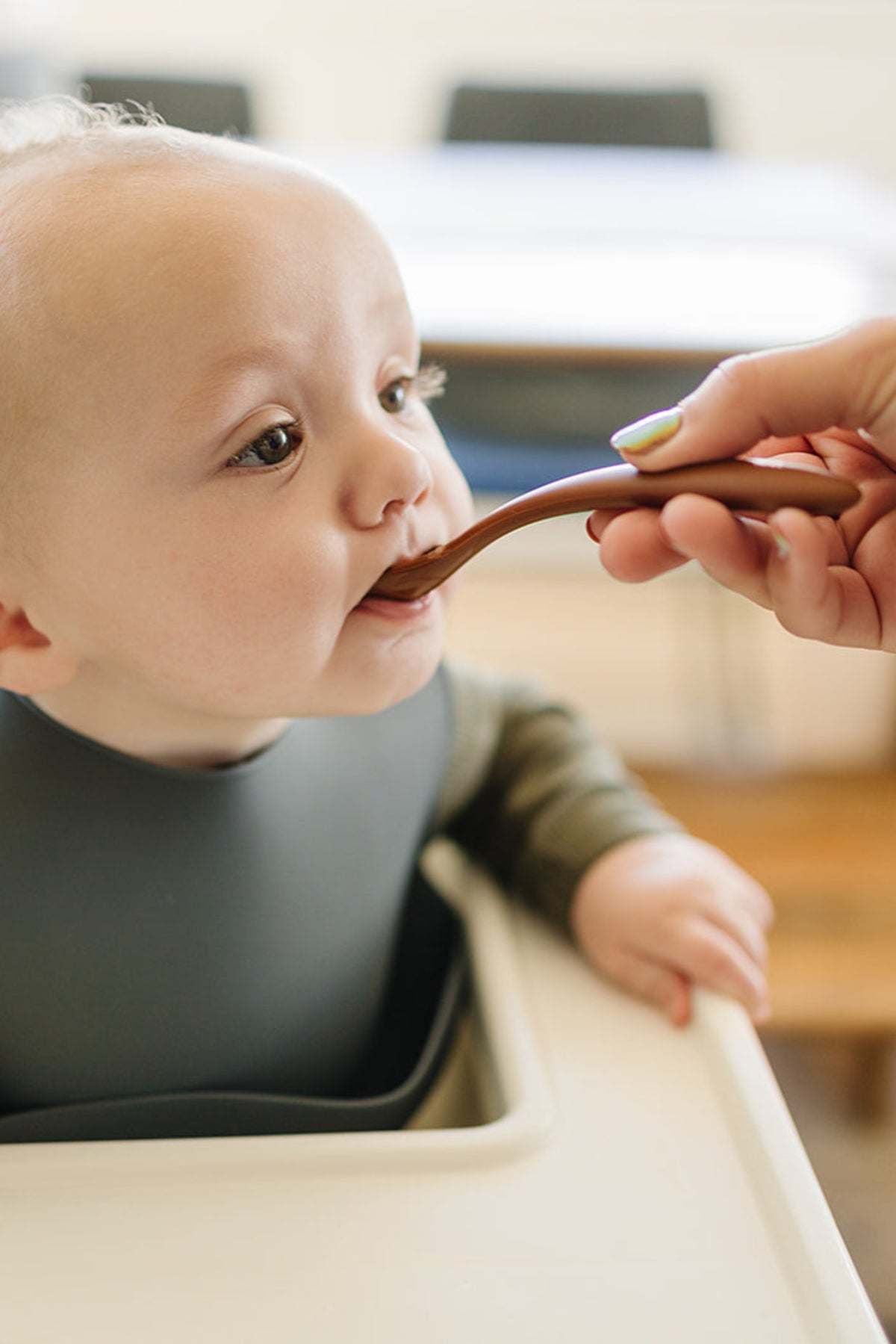 A baby wearing a dark silicone bib is being fed with a brown spoon in a high chair.