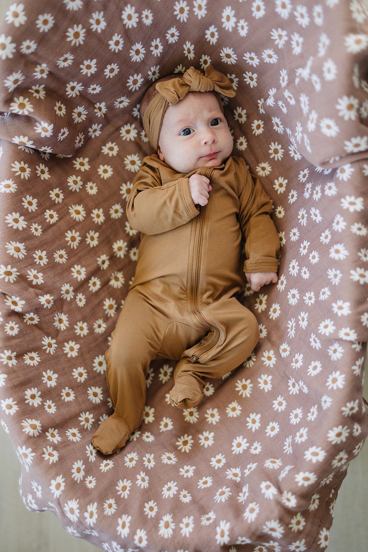 A baby in a mustard footed zipper pajama lays on a floral-patterned blanket, wearing a matching headband.