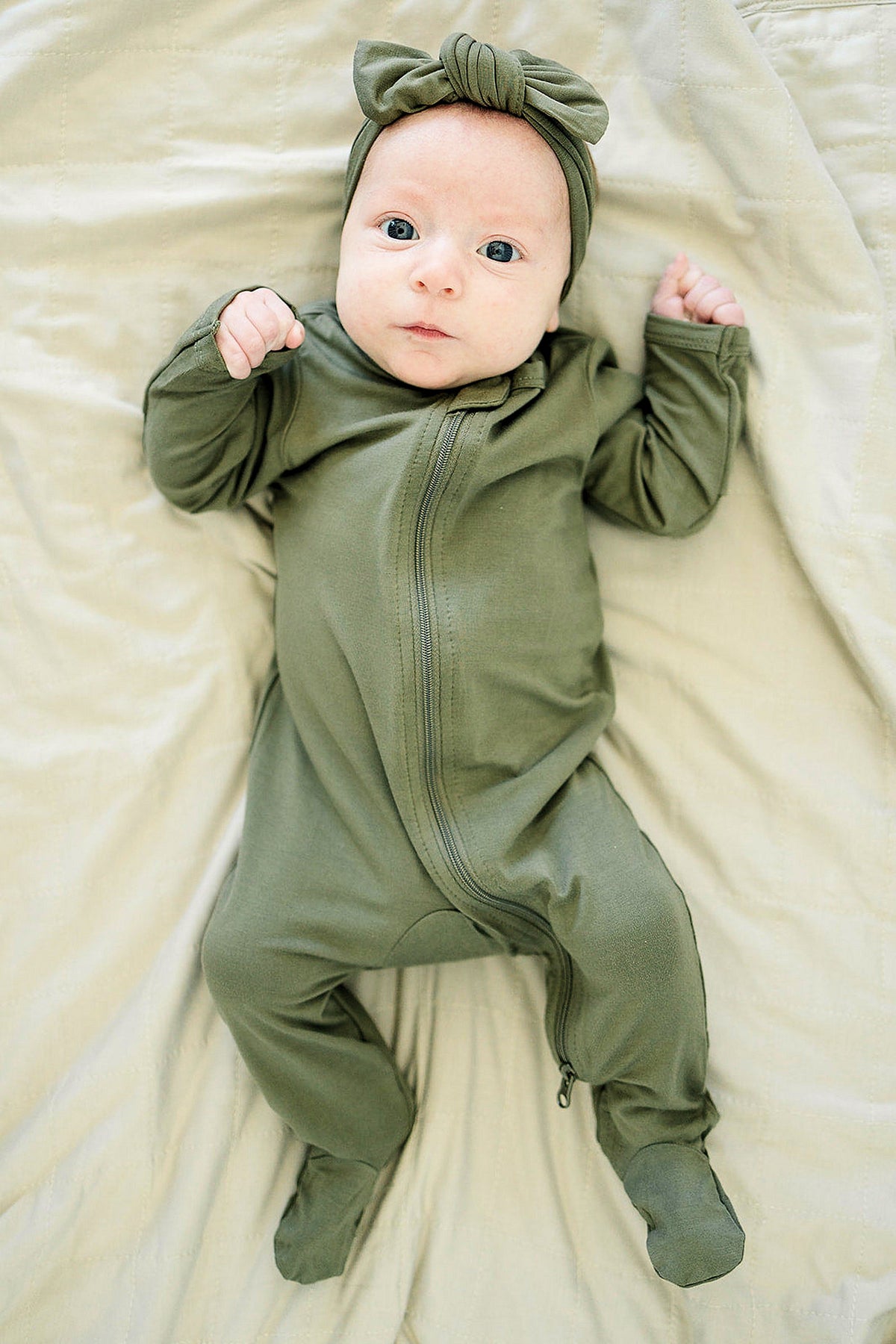 A baby wearing an olive green head wrap and matching zip-up onesie lies on a beige blanket.