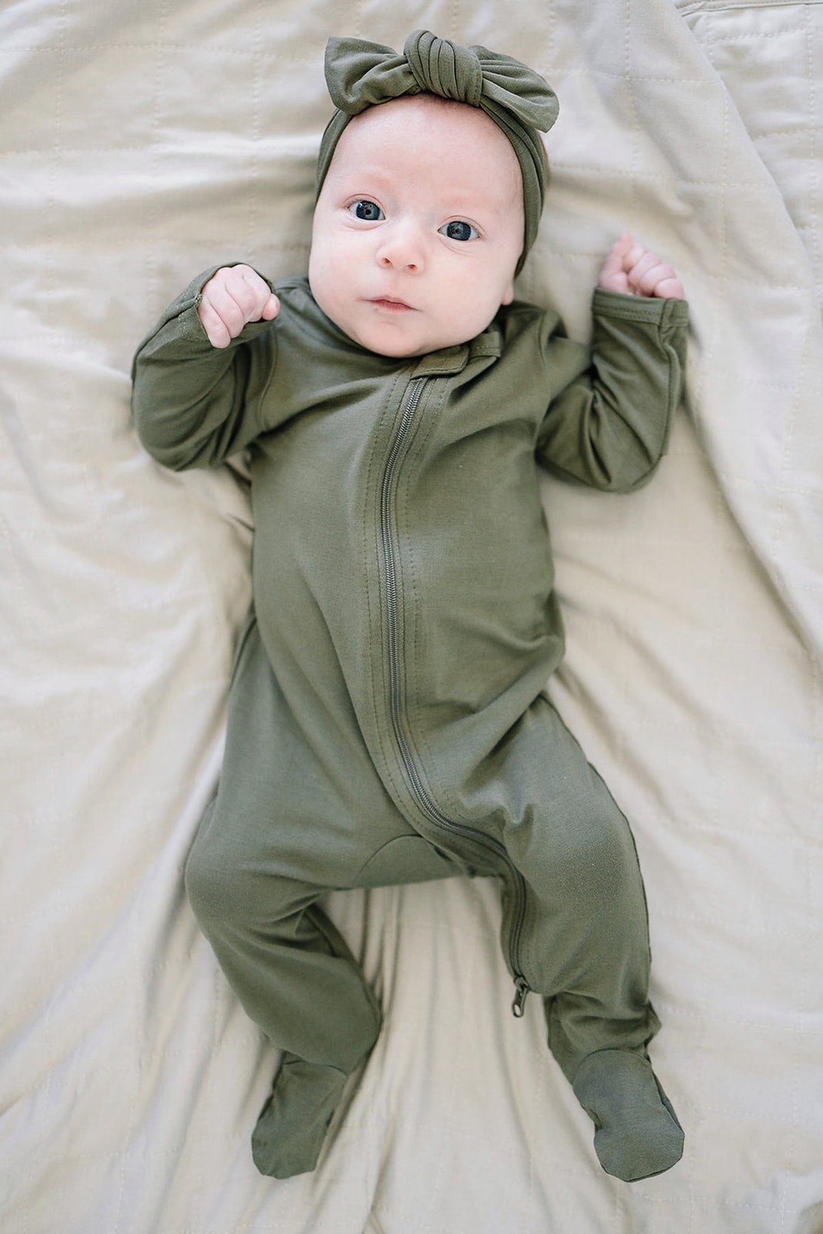 Baby in olive bamboo footed zipper pajama and matching headband, lying on a beige quilted blanket.