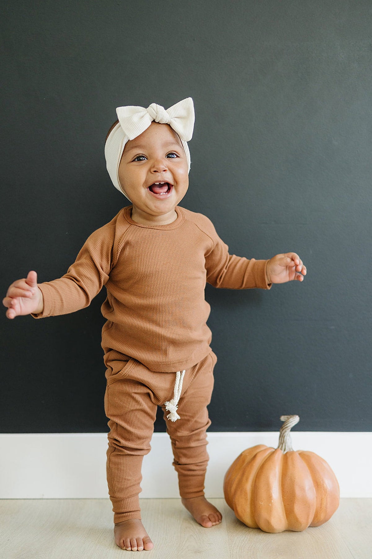 A smiling baby wearing a vanilla ribbed head wrap and brown outfit stands beside a decorative pumpkin.