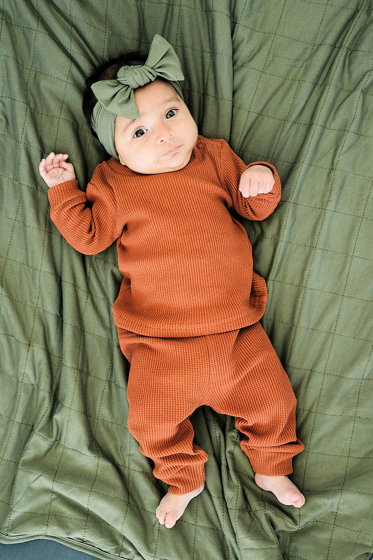 A baby wears an olive head wrap and rust-colored outfit, lying on an olive quilted blanket.