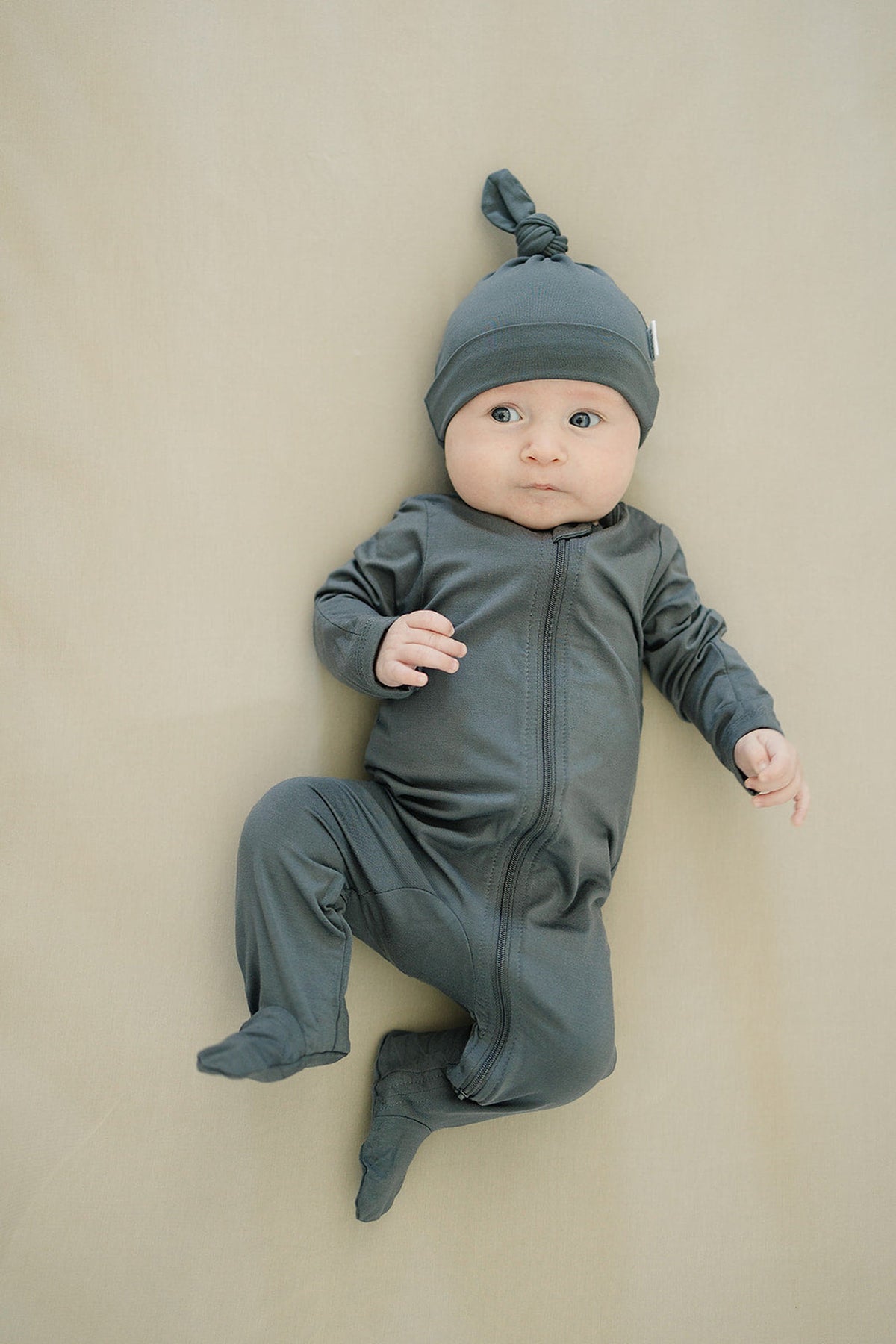 A baby wearing charcoal-colored footed pajamas and a matching knotted hat, lying on a light beige surface.