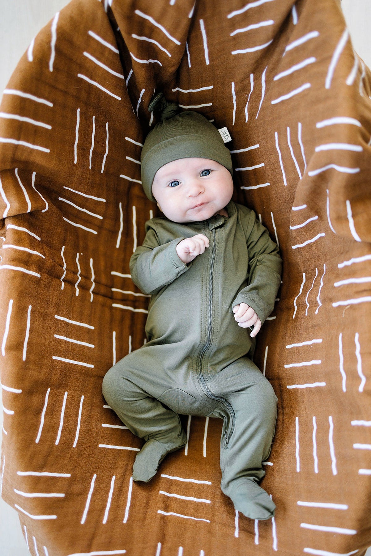 A baby wearing an olive green footed zipper pajama and hat lies on a patterned brown blanket.