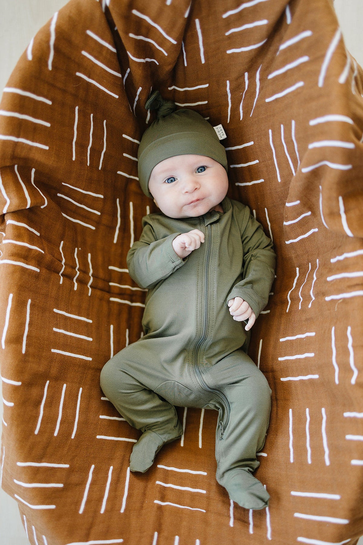 A baby wearing an olive green outfit and knot hat lies on a brown blanket with white patterns.