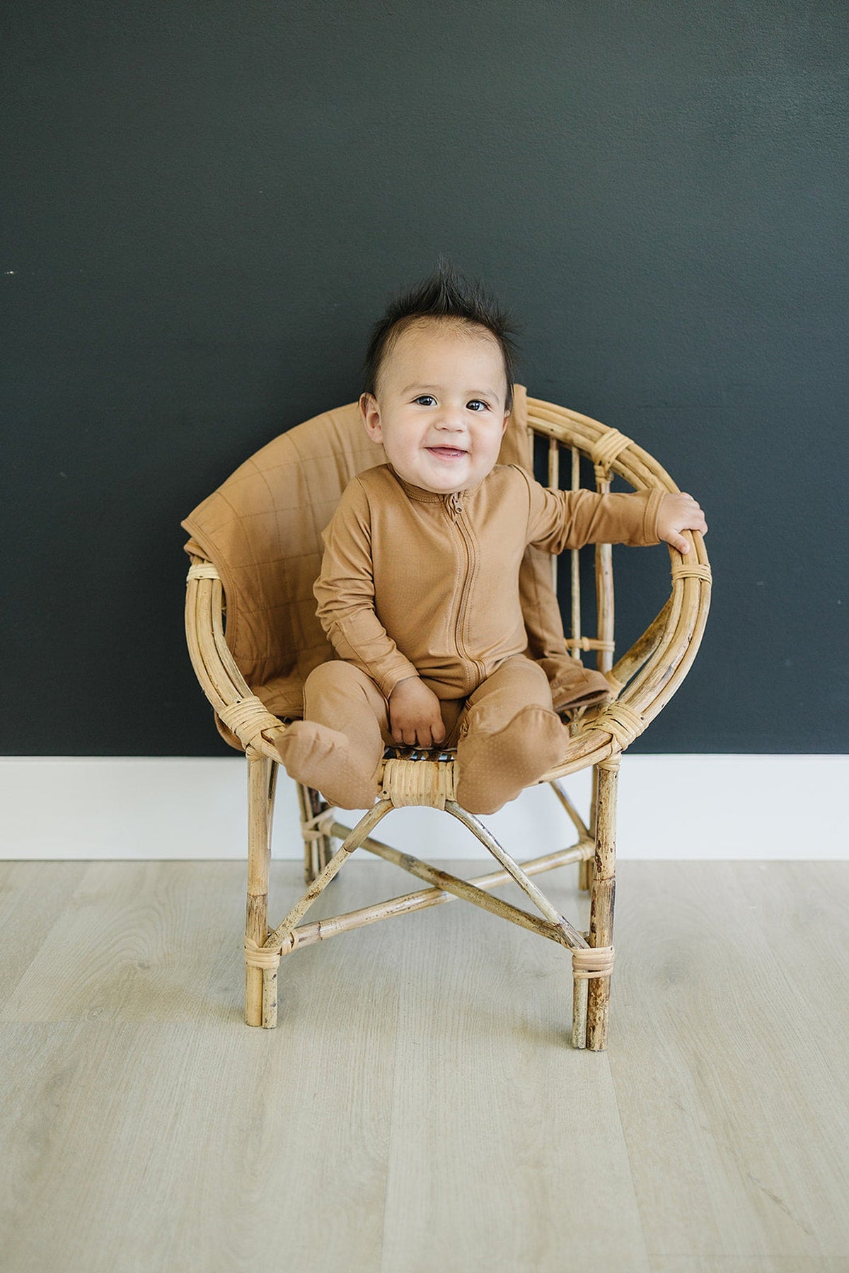 A baby wearing mustard-colored footed pajamas sits in a wicker chair against a dark wall.
