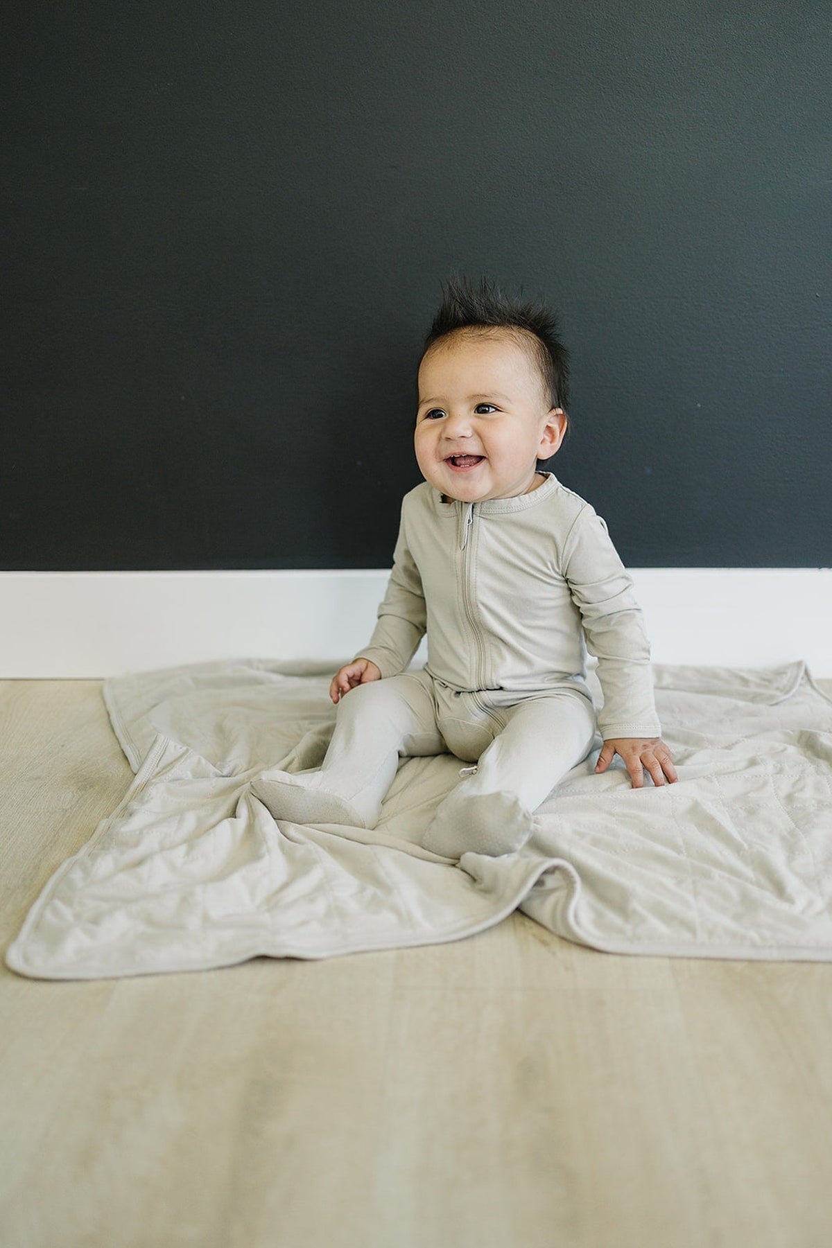 A baby in oatmeal-colored footed pajamas sits on a matching blanket against a dark wall, smiling.
