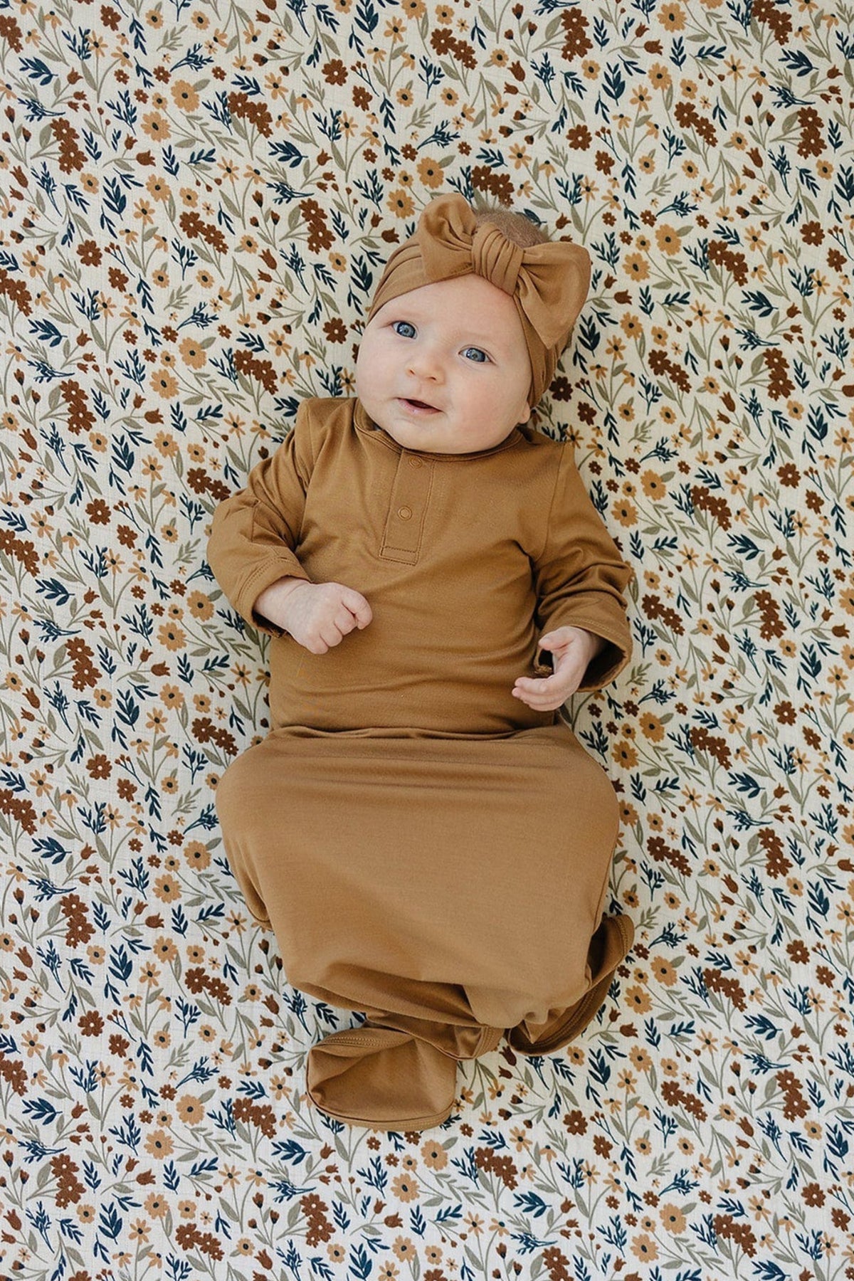A baby in a mustard knot gown and matching headband lies on a floral-patterned blanket.