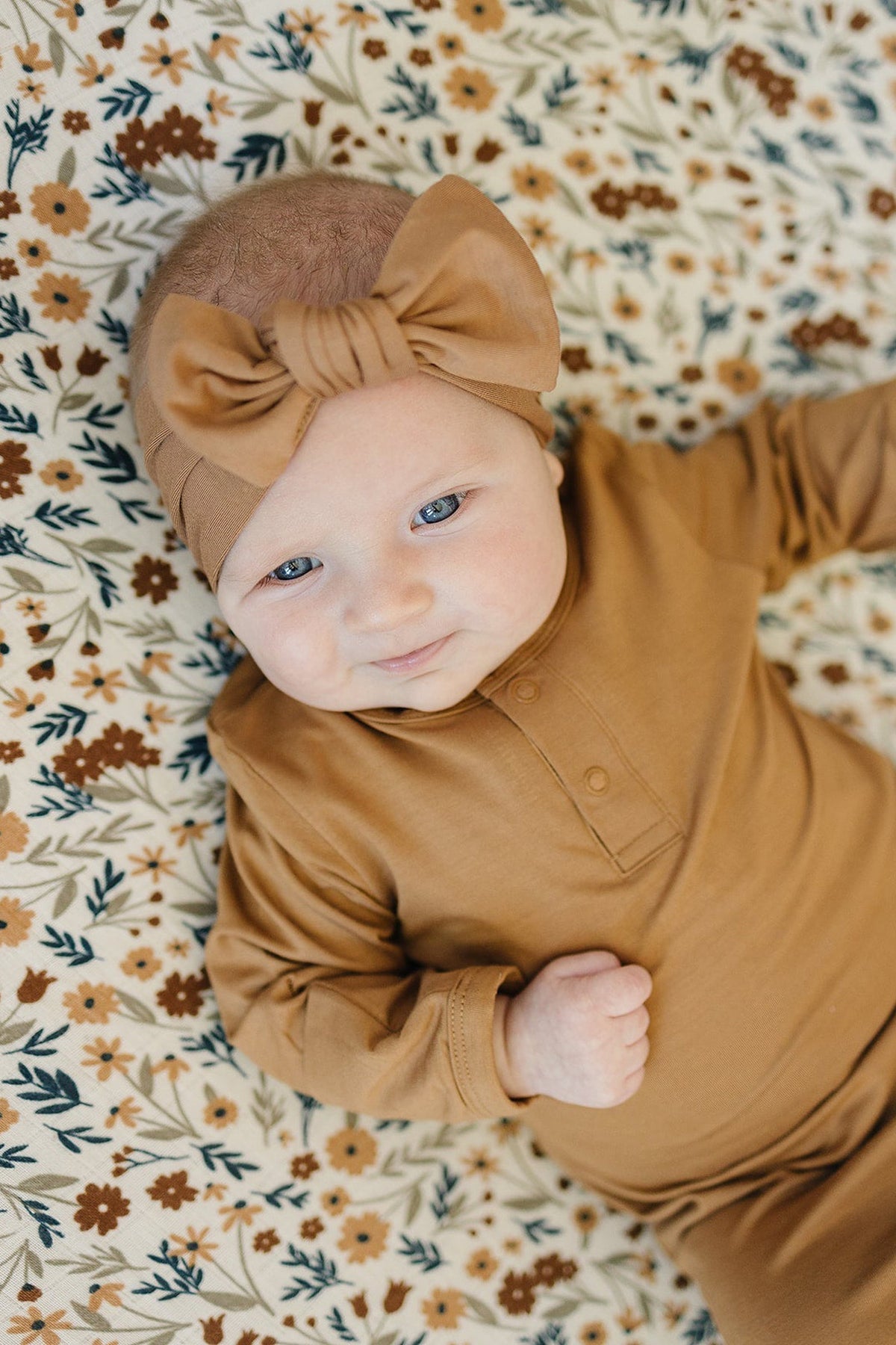 A baby wearing a mustard head wrap and outfit lies on a floral-patterned blanket, smiling softly.