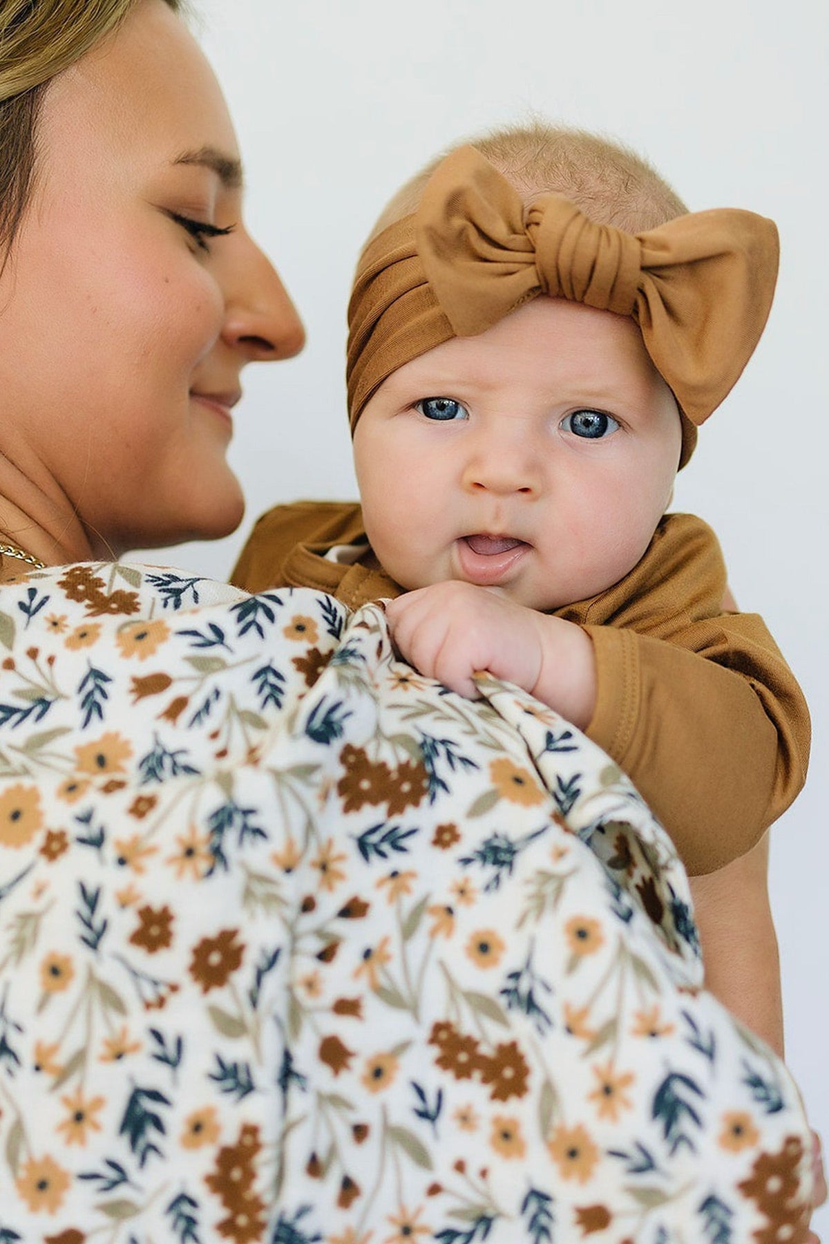 A baby wearing a brown headband is held by a woman, with a floral patterned cloth visible.