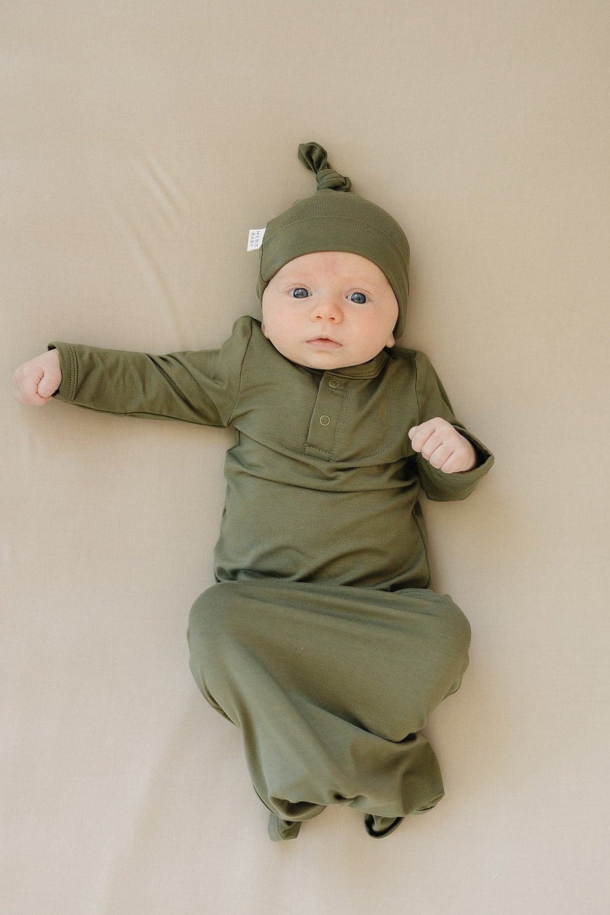 Newborn wearing an olive green knot gown and matching hat, lying on a beige surface.