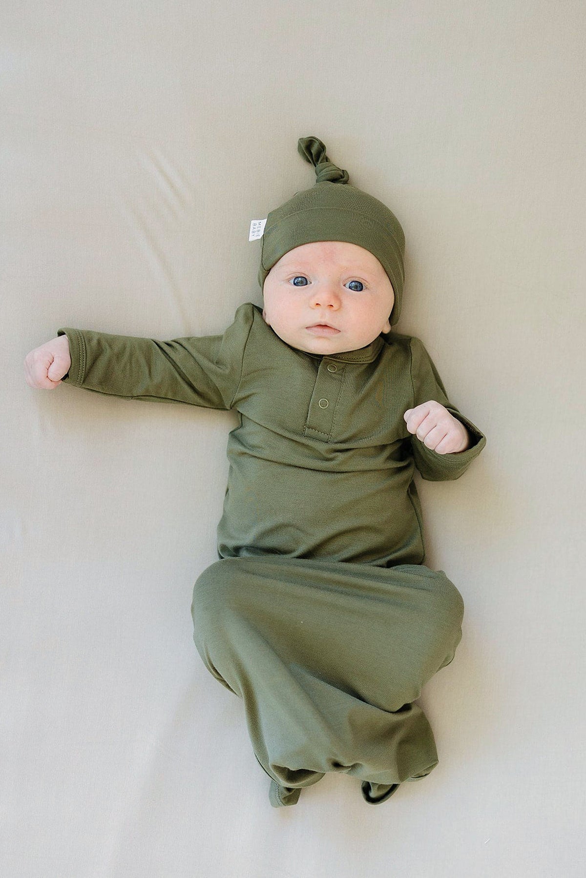 A baby lies on a beige surface wearing an olive green knot gown and matching hat, looking upwards.