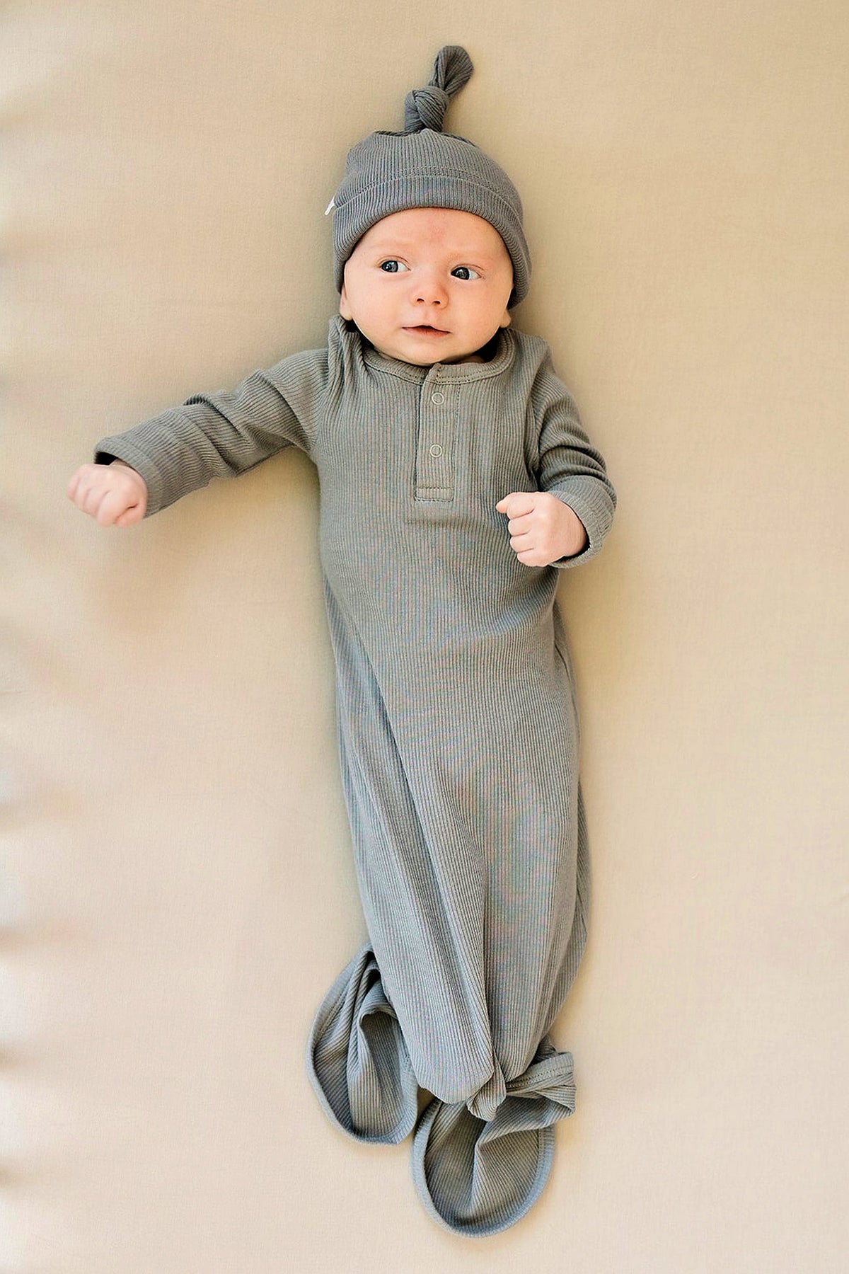 A baby wearing a grey ribbed knot gown and matching hat lies on a beige surface, looking content.