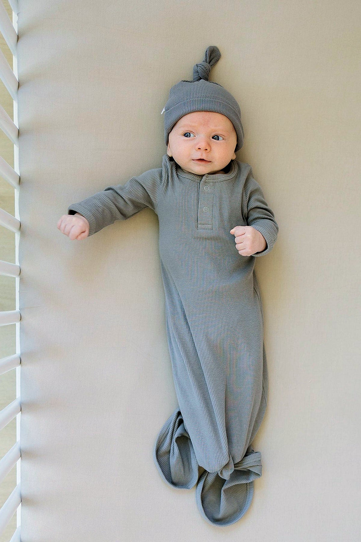 A baby wearing a gray knotted gown and hat lies on a light oatmeal crib sheet in a crib.