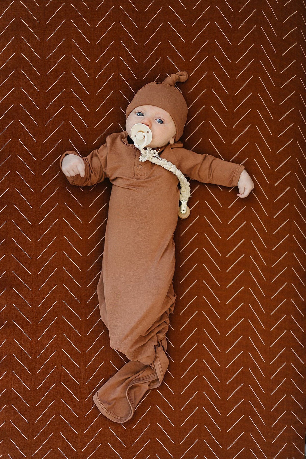A baby in a rust-colored swaddle with a matching hat lies on a patterned muslin crib sheet.