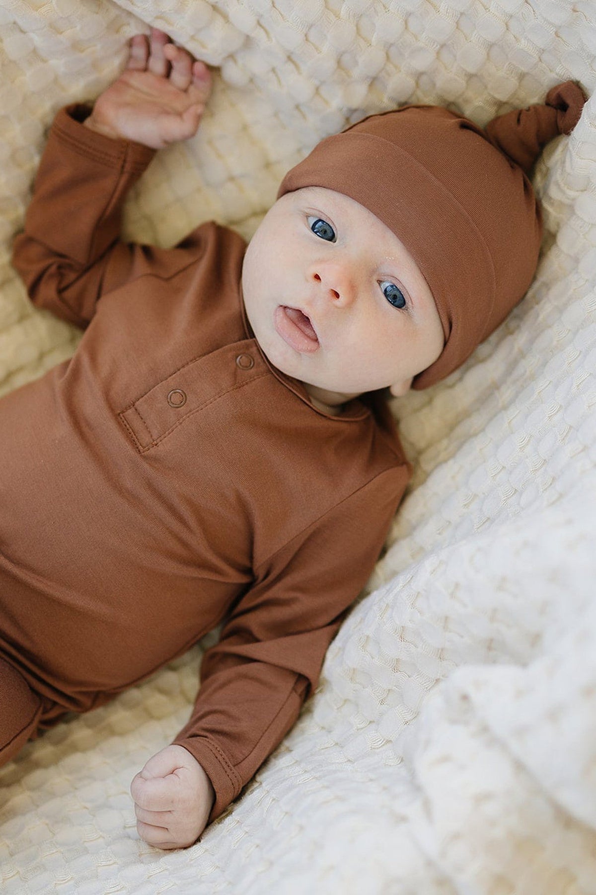 A baby wearing a rust-colored bamboo knot hat and matching outfit lies on a textured white blanket.