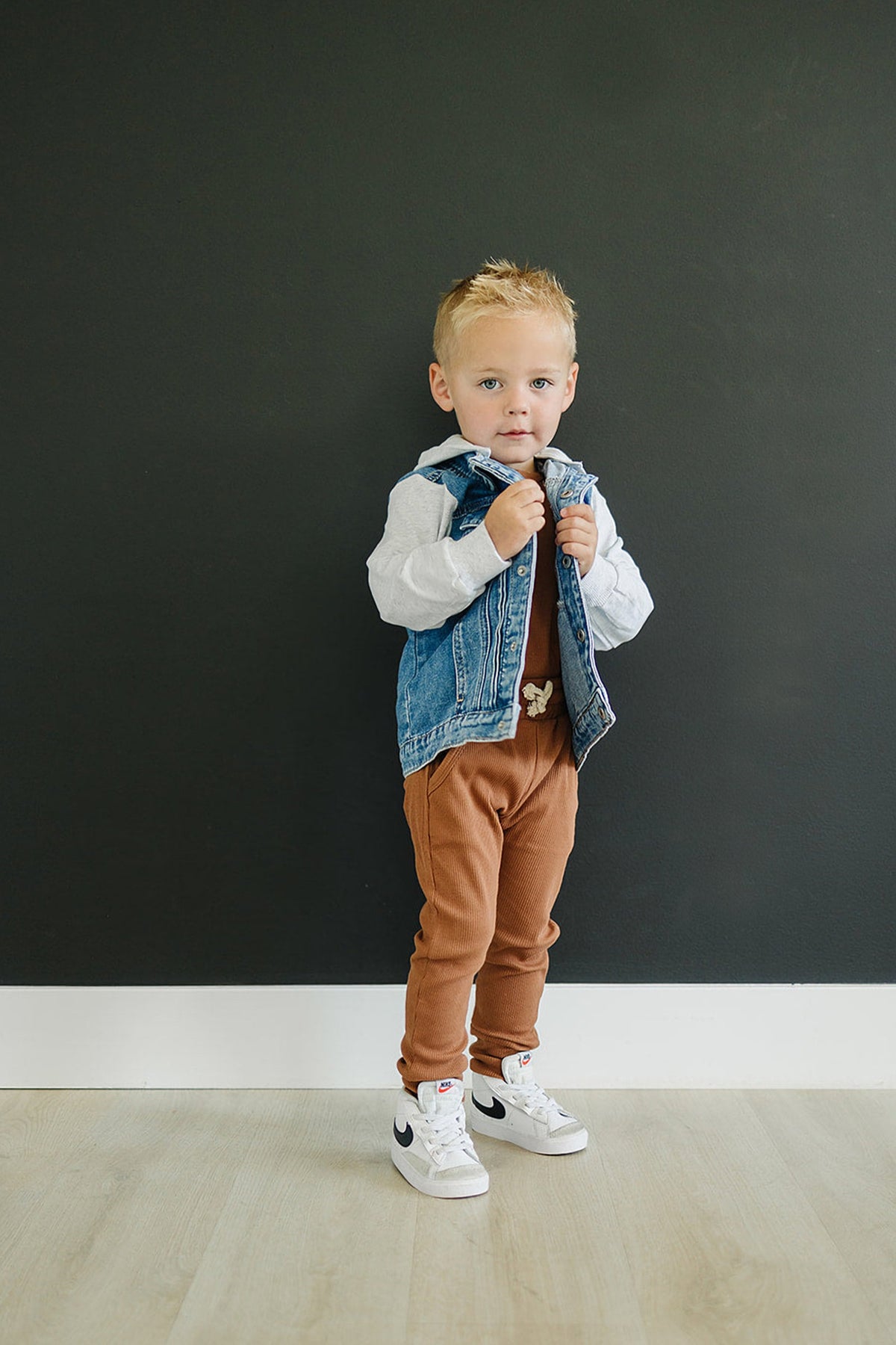 A toddler wears a denim vest with a grey hood over brown pants and white sneakers, standing indoors.