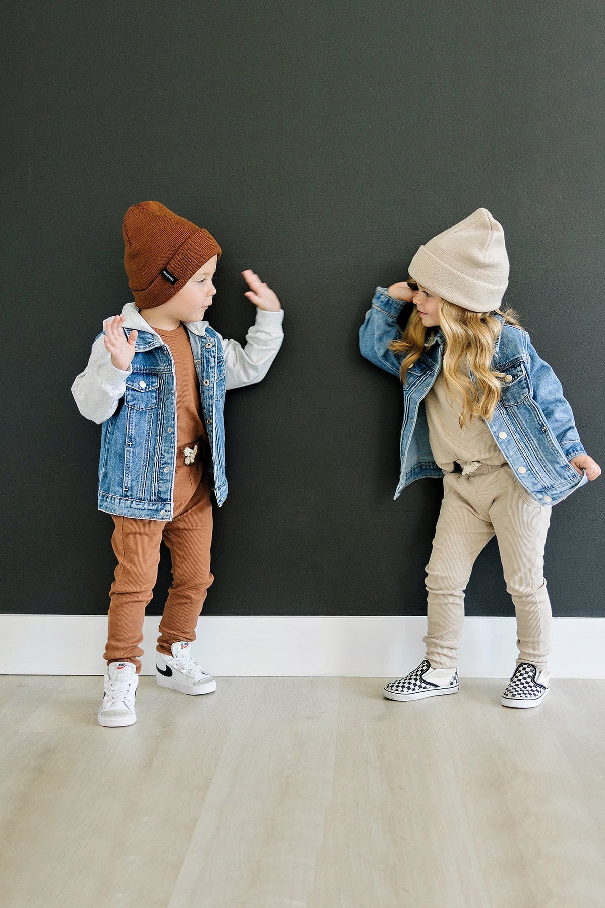 Two children wearing denim jackets and beanies playfully pose against a dark wall on a wooden floor.