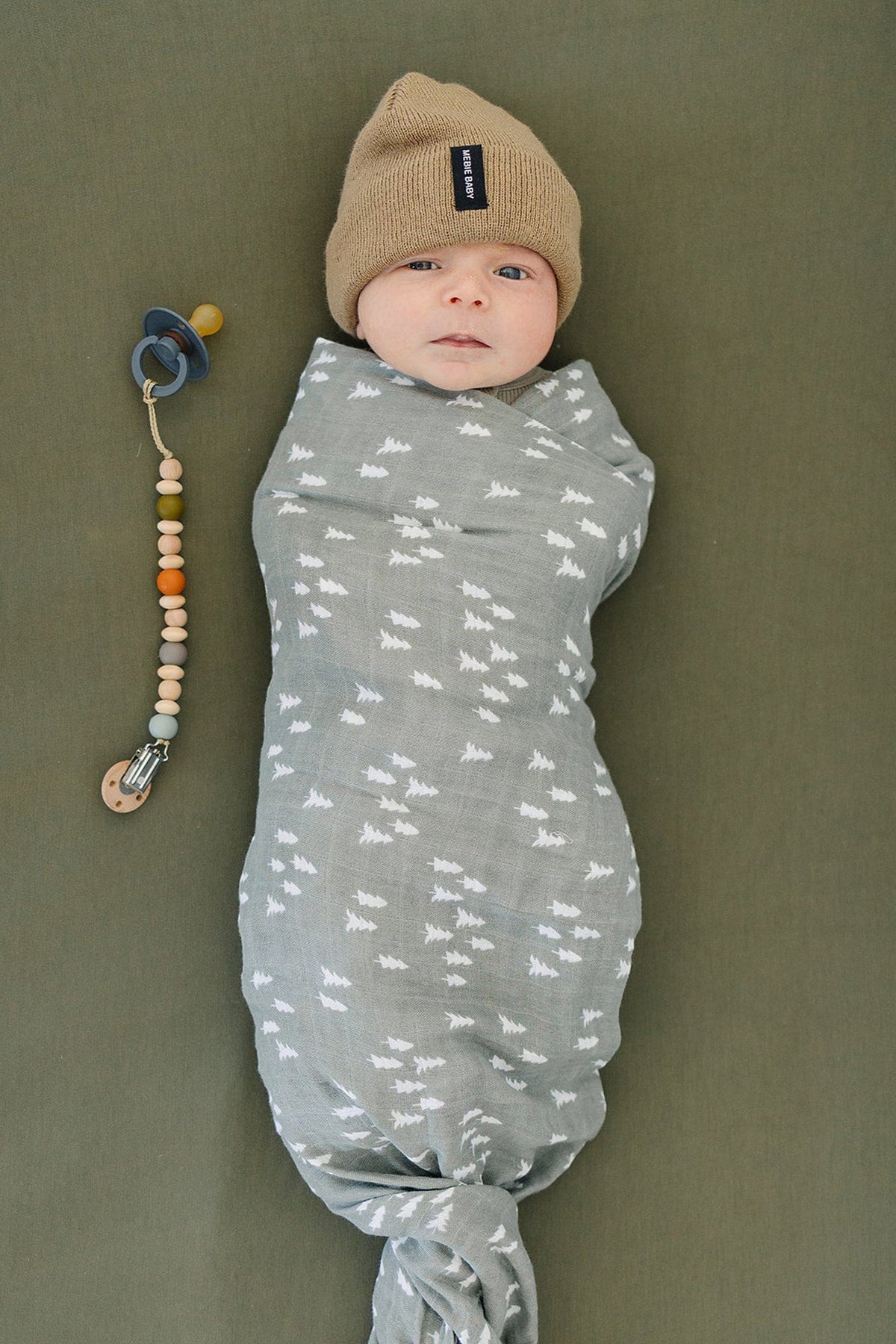 A baby wrapped in a gray muslin swaddle with pine patterns, wearing a brown knit hat beside a pacifier.