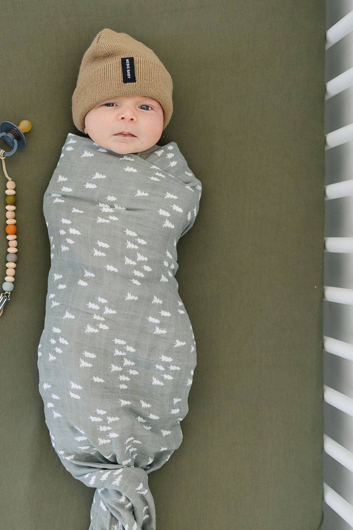 A baby swaddled in a gray blanket with white patterns lies on an olive crib sheet, wearing a tan beanie.