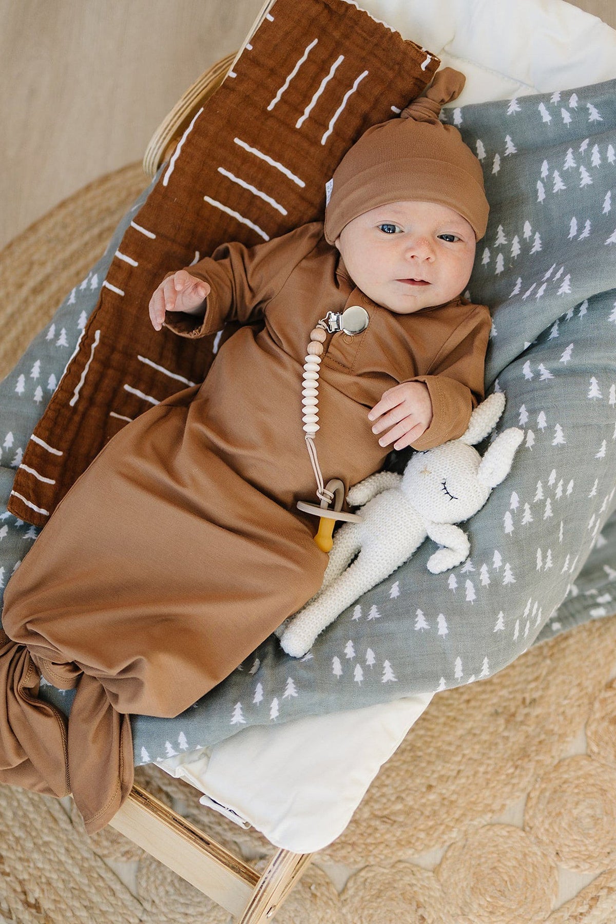 A baby in a brown knot hat and gown lies on patterned blankets with a soft toy rabbit.