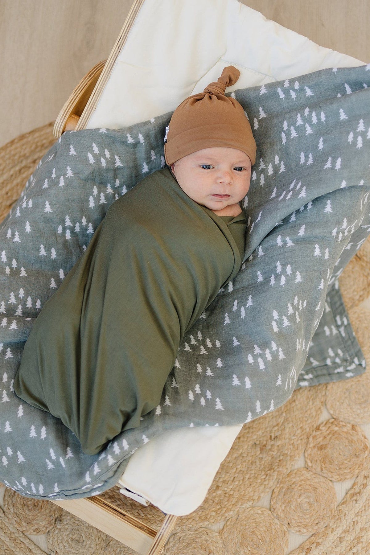 Newborn wearing a mustard knot hat, swaddled in green fabric, lying on a tree-patterned blanket.