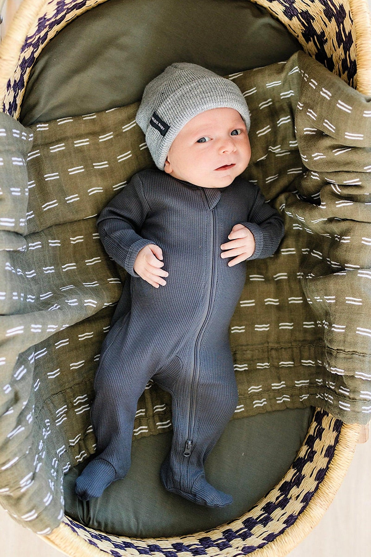 A baby in a charcoal ribbed zipper footed pajama lies in a patterned basket, wearing a gray beanie.