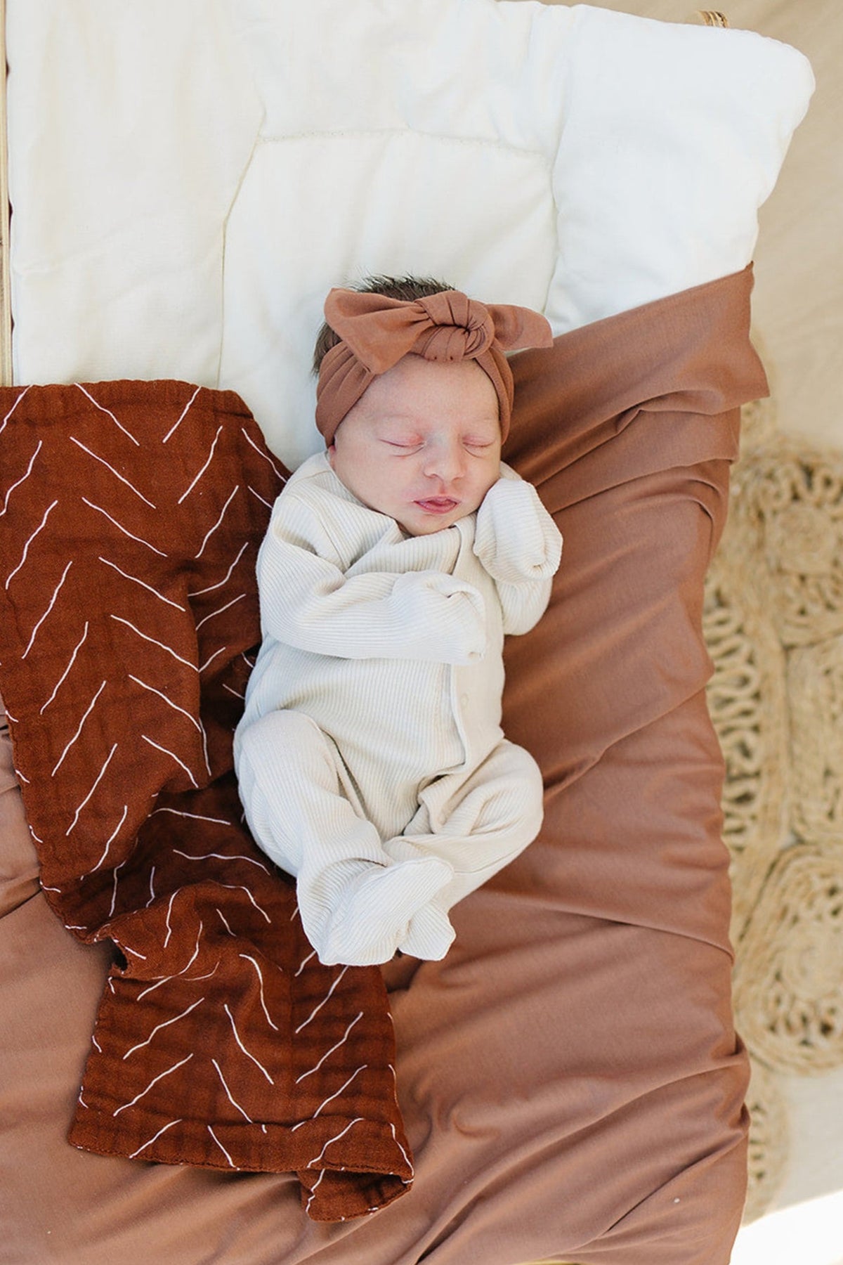 Newborn in cream ribbed pajamas and brown headband, lying on a brown and white blanket.