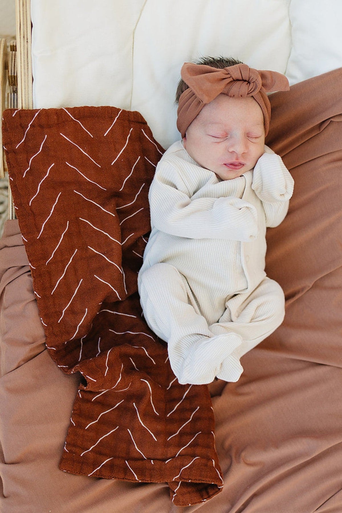 A newborn baby sleeps on a rust-colored, patterned burp cloth, wearing a matching headband and light outfit.