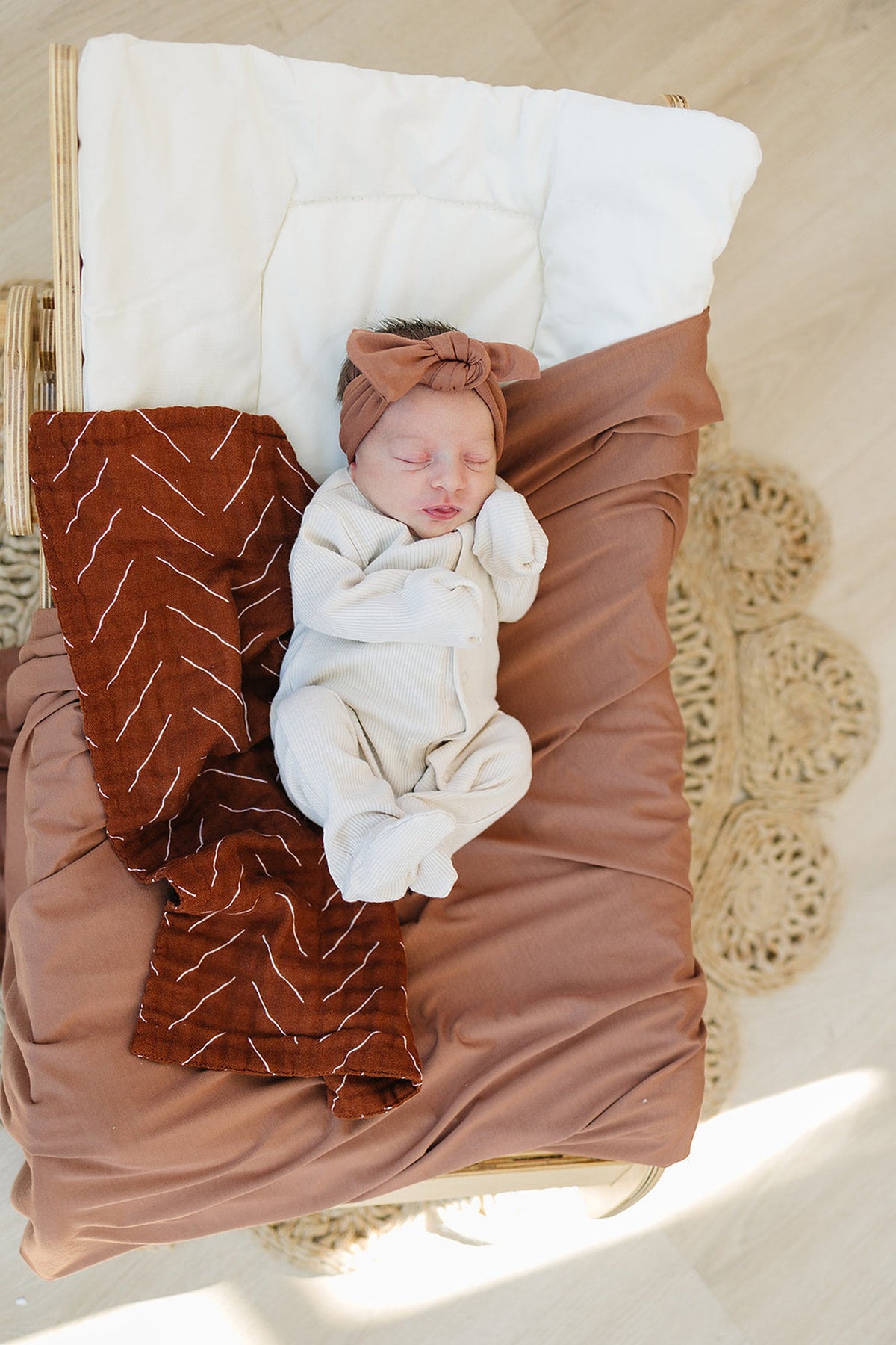 A newborn in a cream outfit and rust head wrap is swaddled in a rust-colored bamboo blanket.