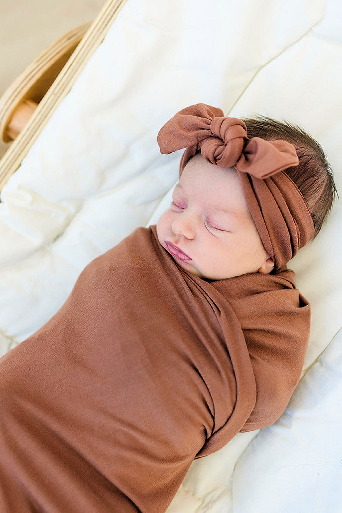 A sleeping baby wrapped in a rust-colored bamboo swaddle with a matching head wrap, resting on a soft surface.
