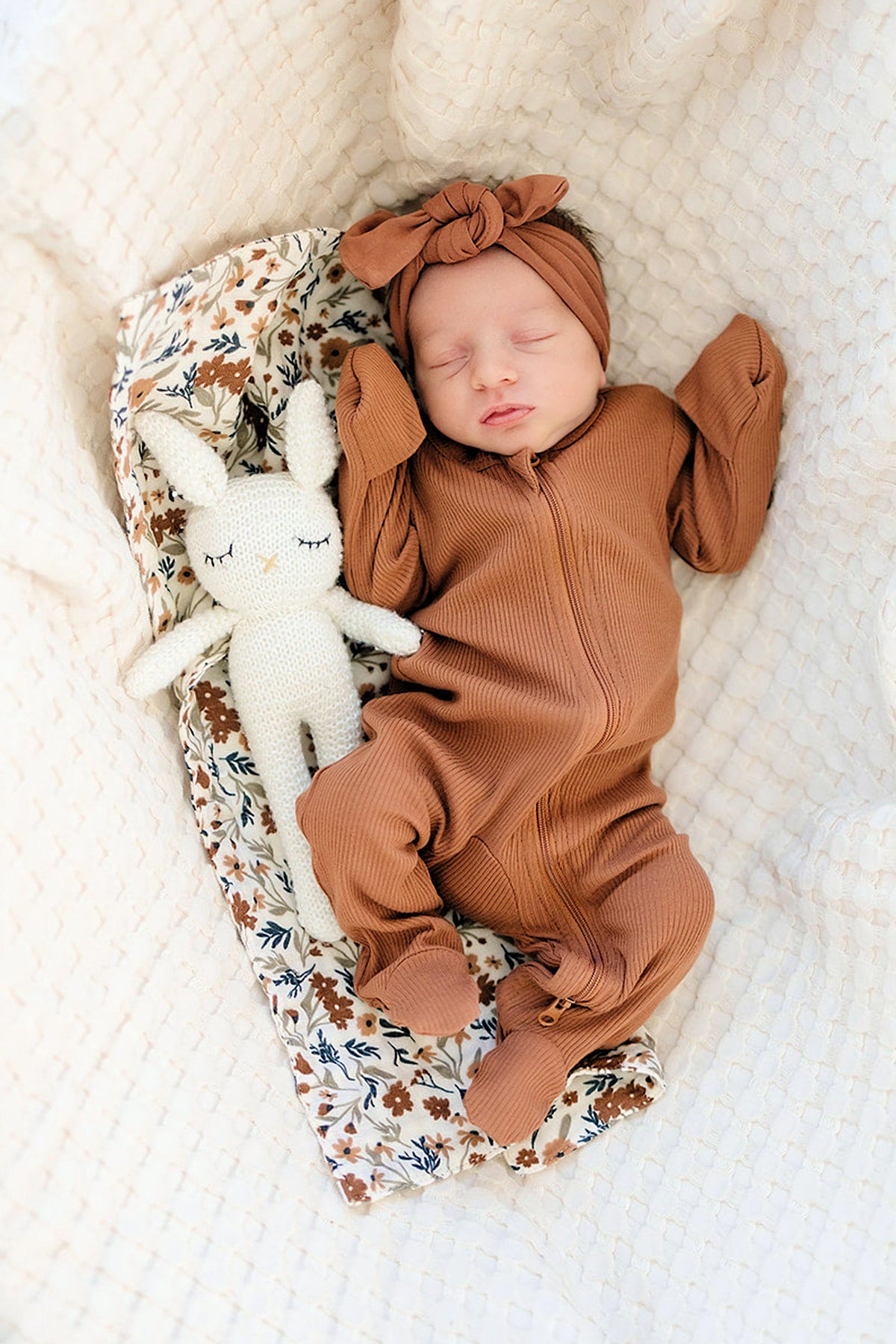 A baby in a brown outfit and headband sleeps on a floral-patterned cloth next to a knitted toy.