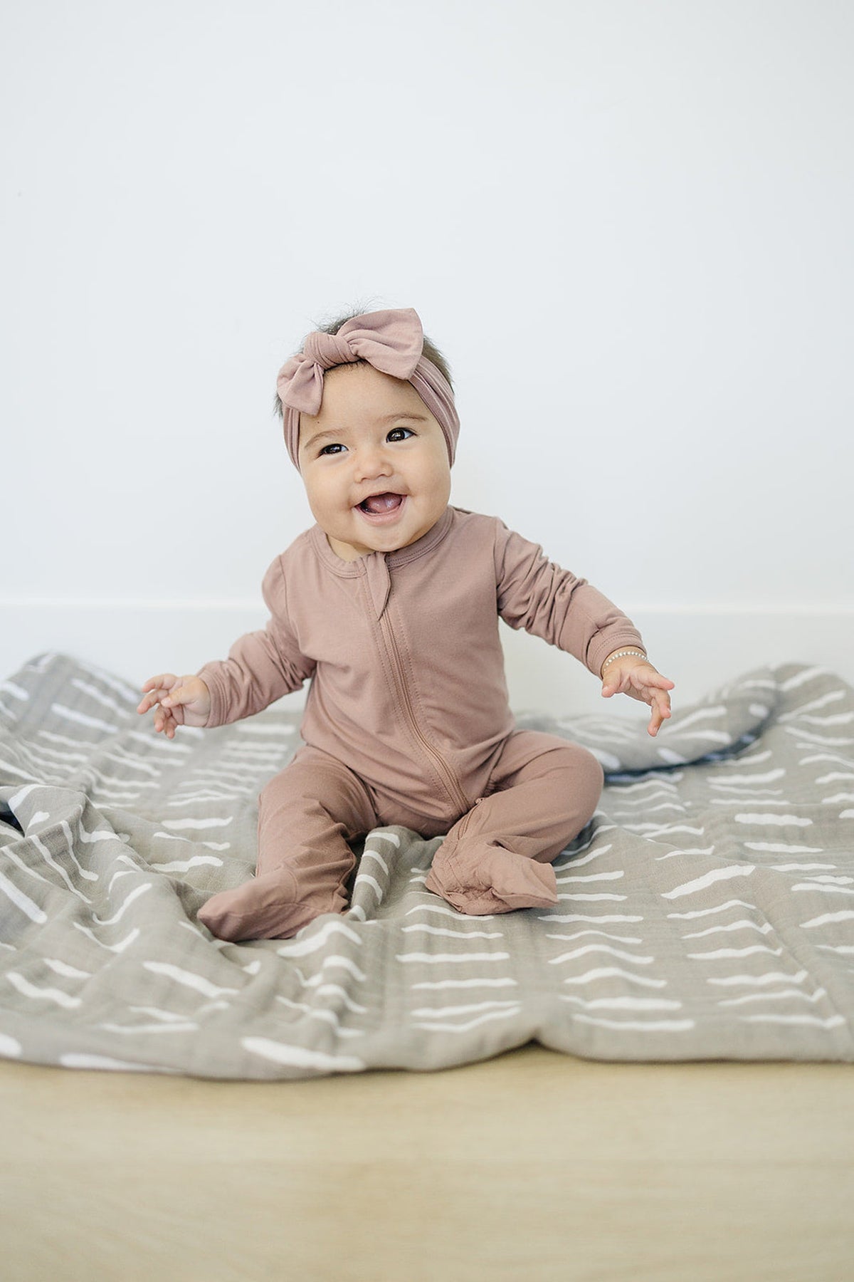 A baby wearing dusty rose footed pajamas and headband sits on a patterned gray blanket, smiling happily.